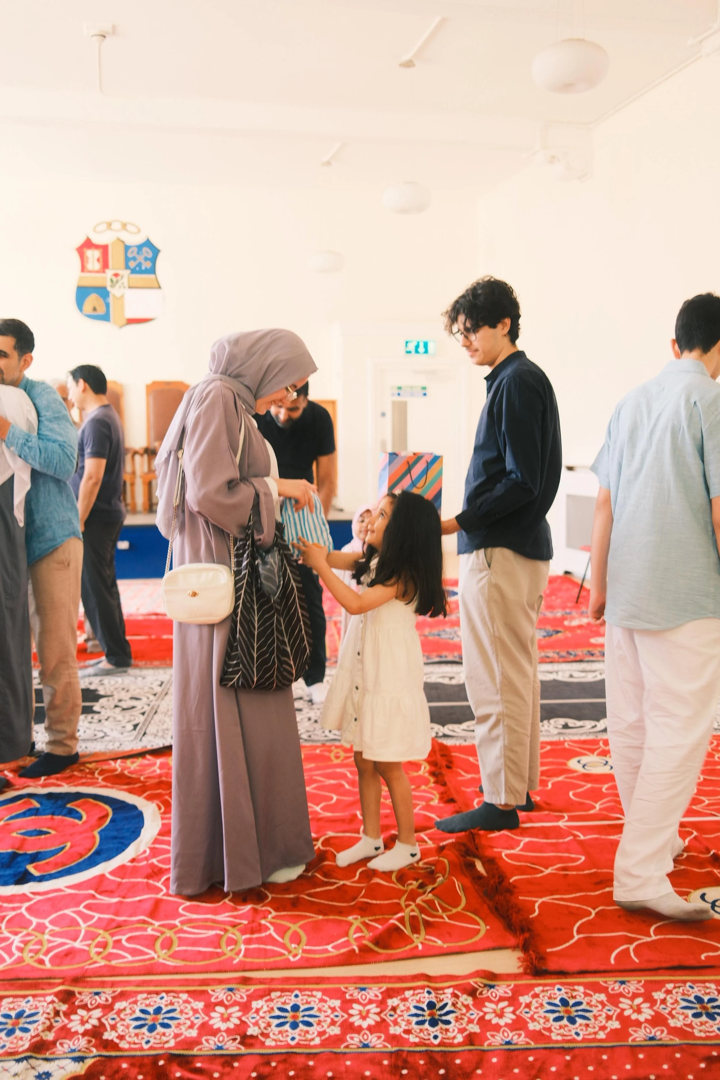 People gathered in a room with a red and black patterned carpet, engaging in a social or religious event. Women and children are interacting, with some women wearing hijabs, and small gift bags are visible. In the background, there are multiple peopl