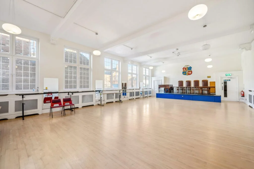 Eastbourne Town Hall, a large, bright room with high ceilings, multiple large windows, light wood flooring, and a small elevated stage with chairs and a crest on the wall.