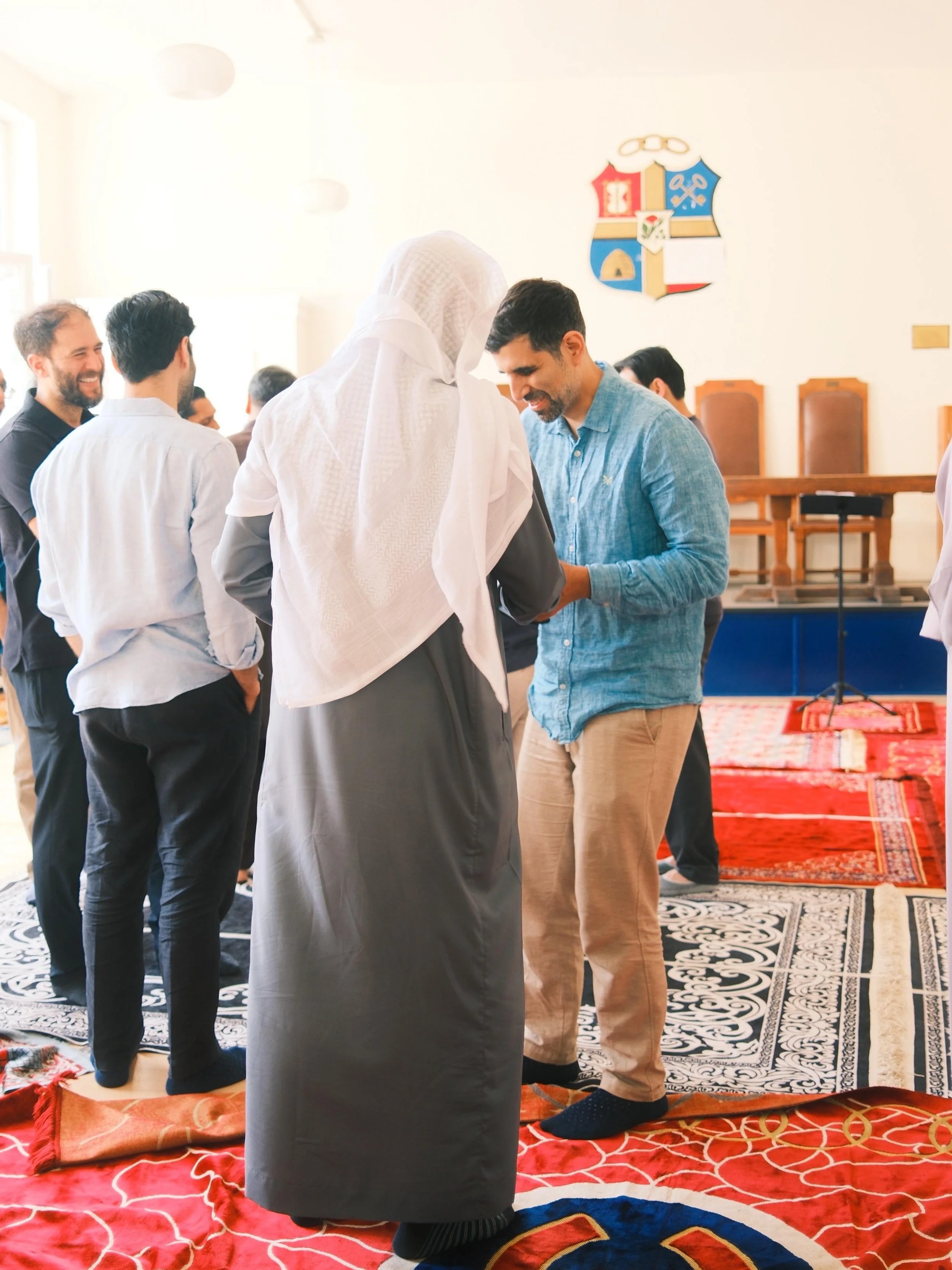 A group of men gathered in a mosque, with one man wearing traditional Islamic attire and the others in casual clothes, standing and talking on prayer rugs.