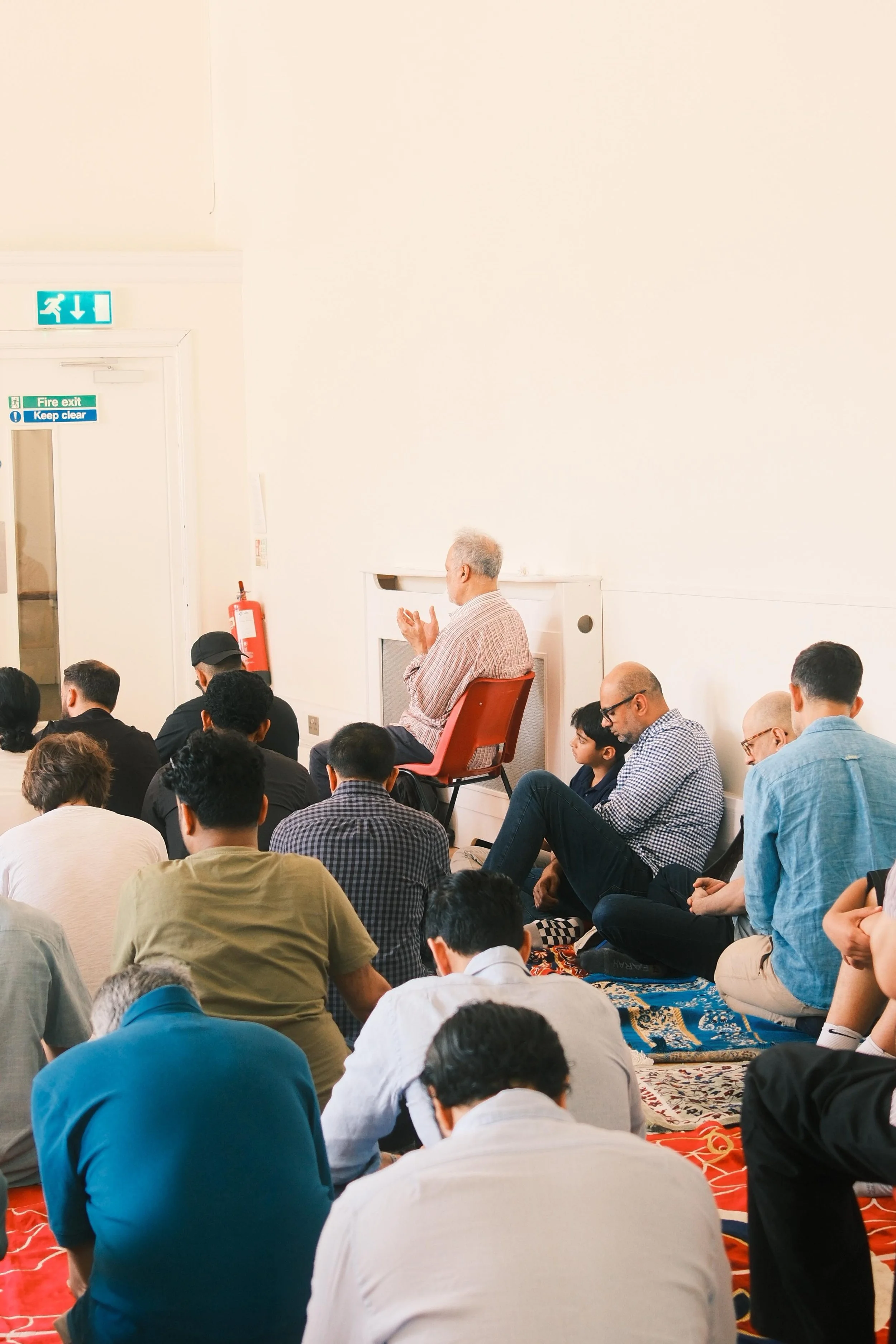 A man sitting on a chair, praying or meditating during a congregation inside a mosque. Several attendees are sitting on the carpeted floor around him, with some looking down or closing their eyes.
