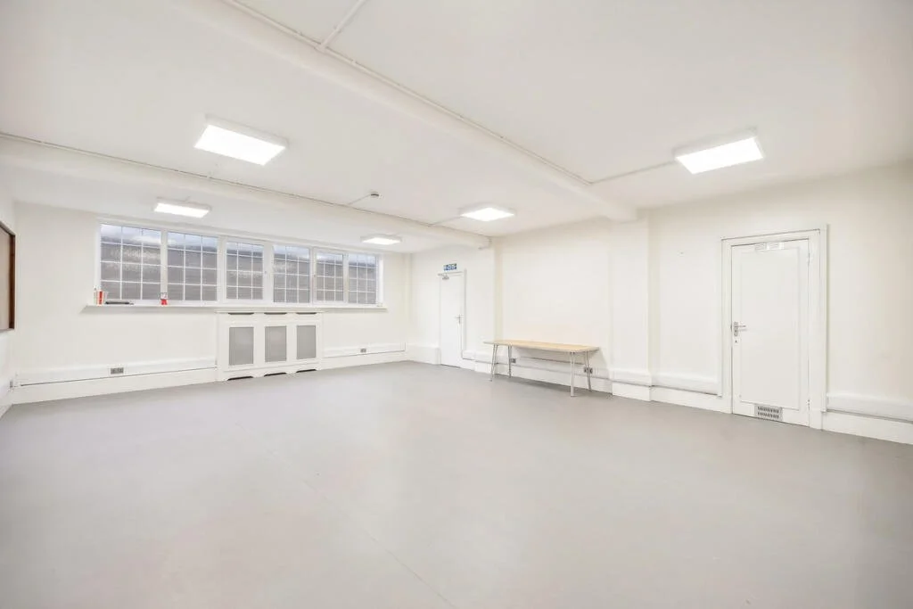 Empty white room with large windows, overhead fluorescent lights, a small table, and doors