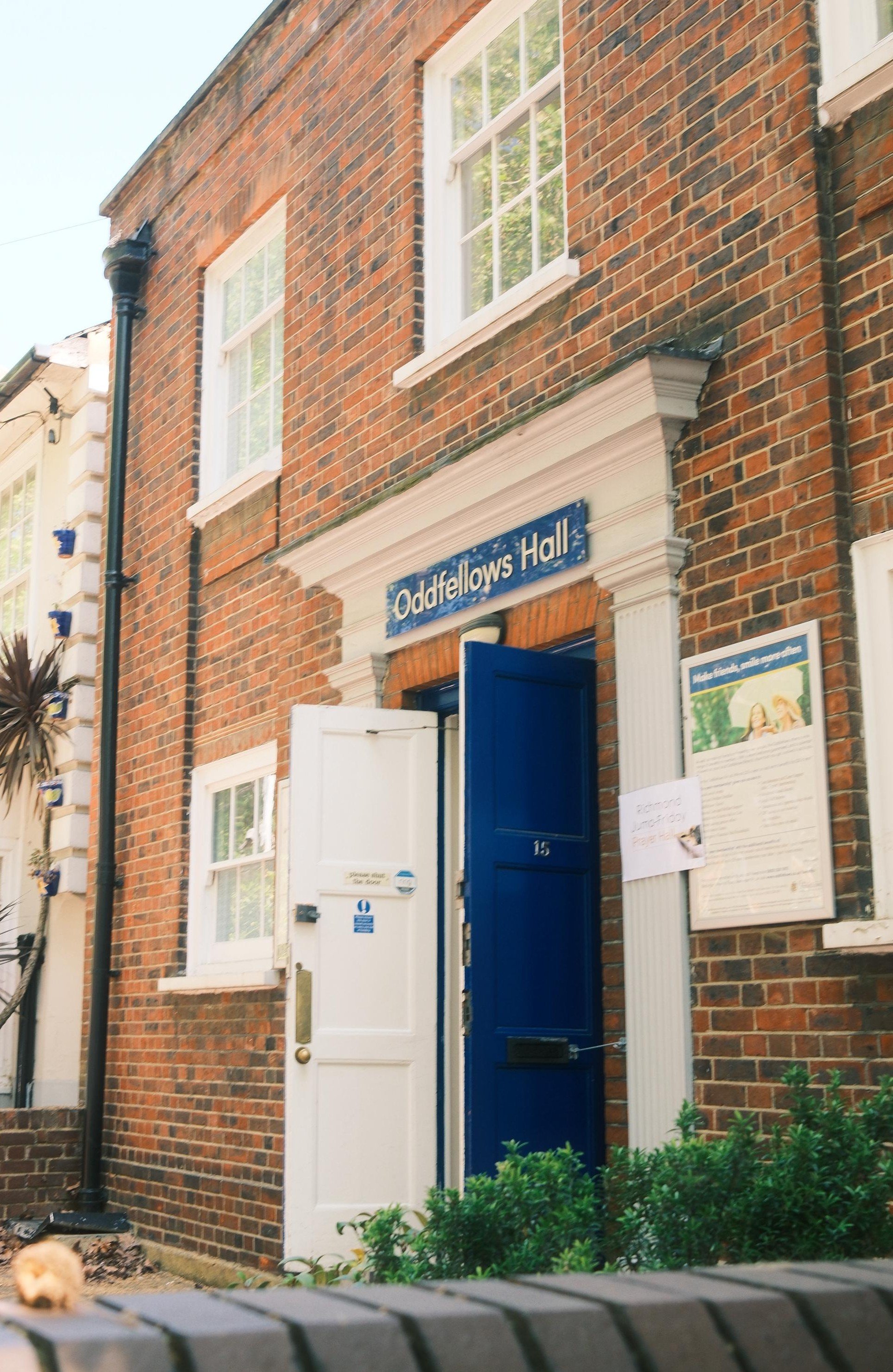 Brick building with white-framed windows, a blue door with the number 15, and a sign reading "Oddfellows Hall". There is a white door to the left of the blue door, and a bulletin board with notices to the right. Bushes and greenery are in front of the building.