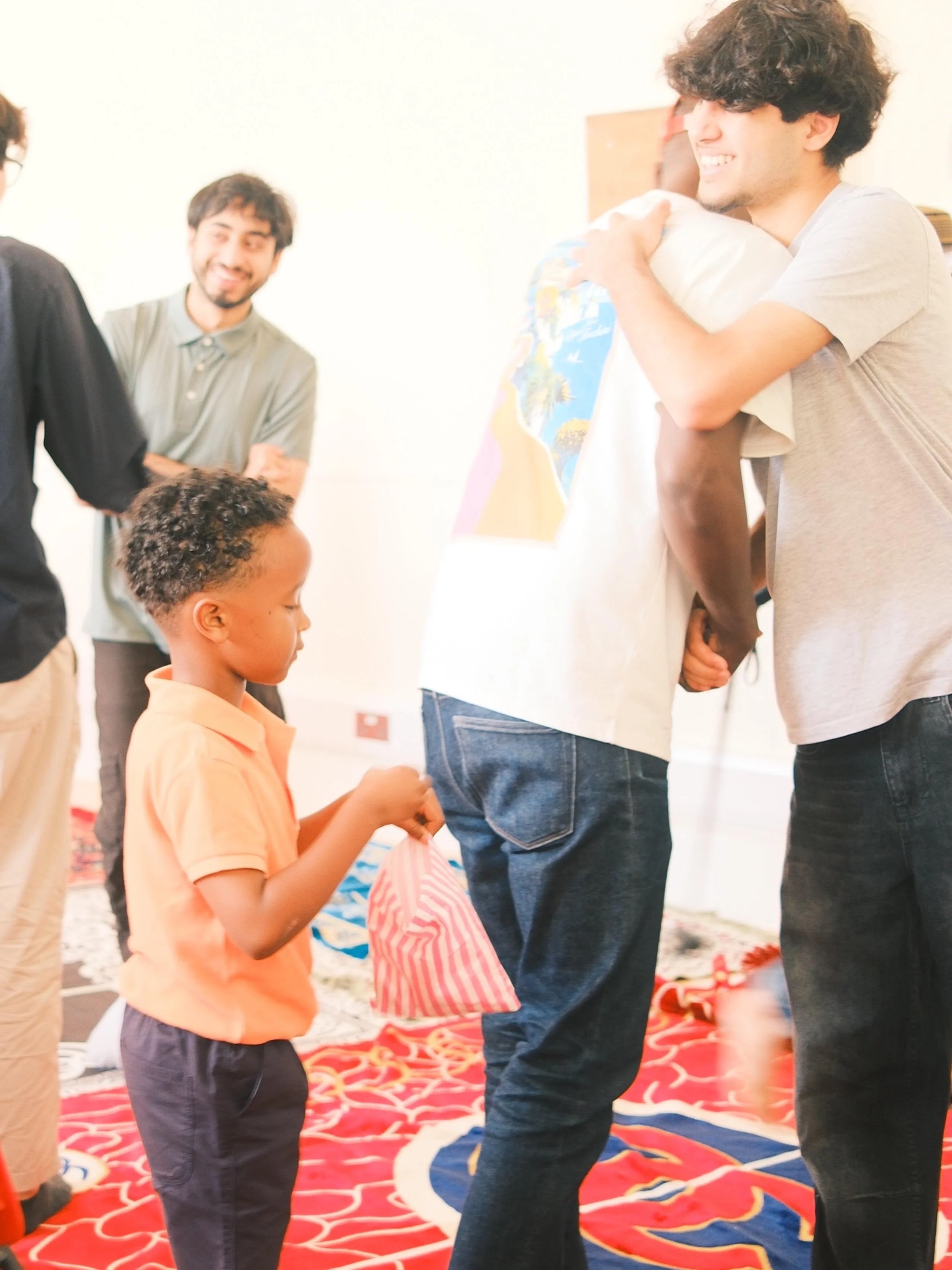 Young boy receiving a gift from a smiling adult, with several other smiling adults nearby in an indoor setting with a colorful carpet.