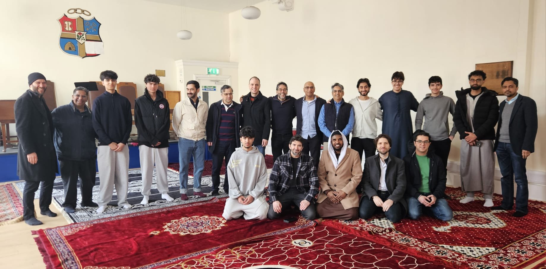 Group of men and boys in a room, some seated on the floor, others standing, posing for a photo on a patterned carpet with a plain, light-colored wall in the background.