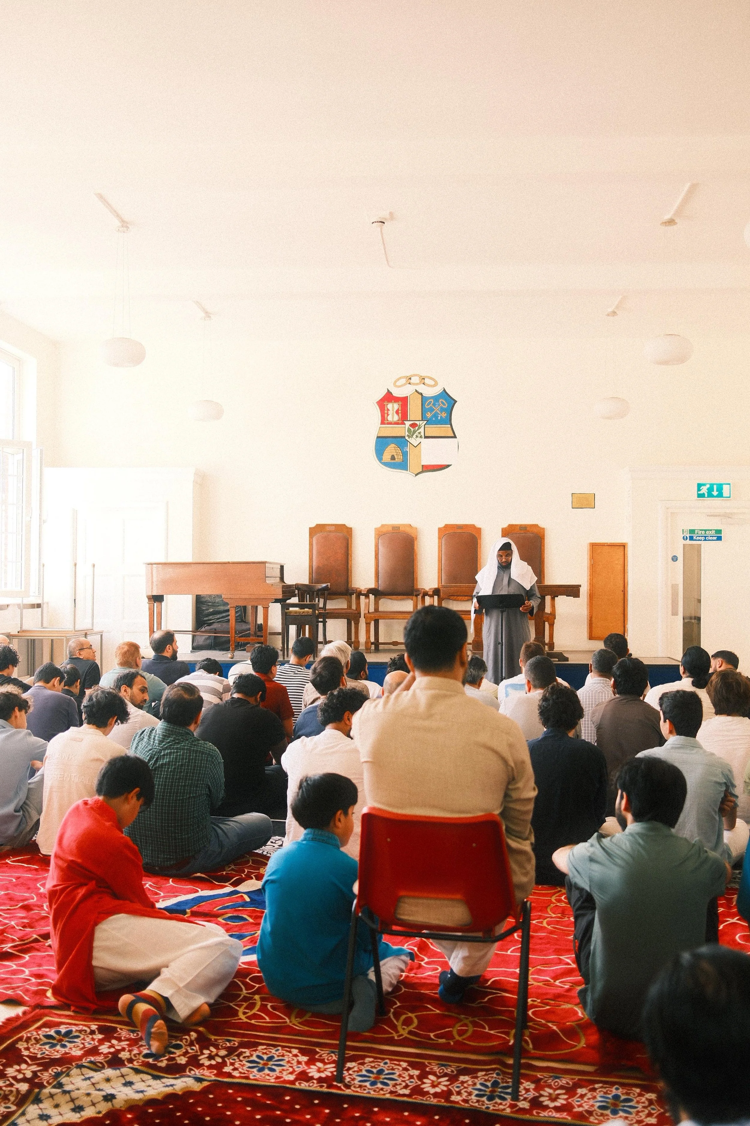 A group of people sitting on a red patterned carpet in a hall, listening to a person in academic dress speaking at a podium. The hall has a high ceiling with ceiling lights, a coat of arms on the wall, and emergency exit signs.