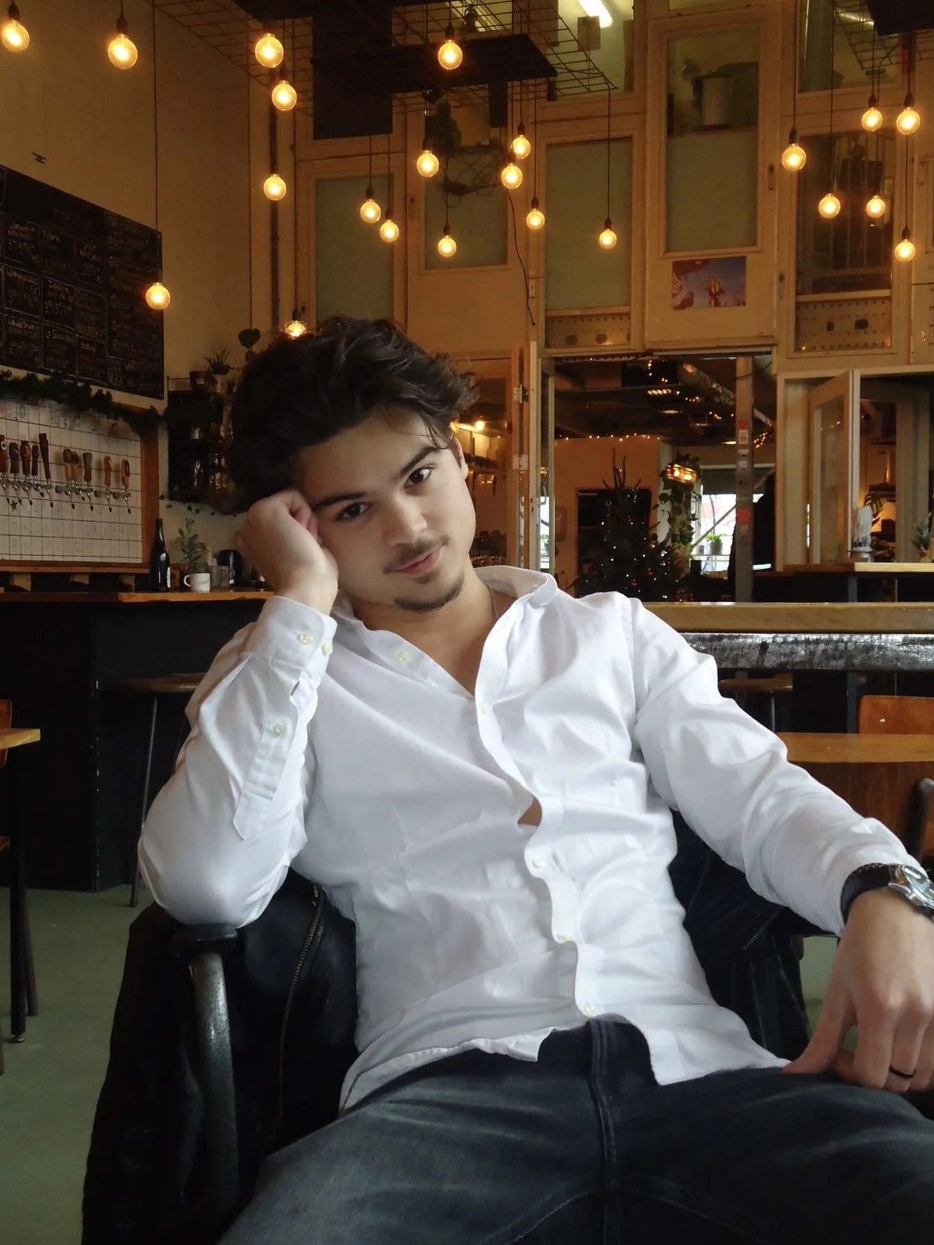 Een jonge man met donker haar en een lichte huidskleur zit in een gezellige kroeg of café, gekleed in een witte overhemd met de knopen open, met een ontspannen houding en een lichte glimlach. Op de achtergrond hangen decoratieve lichtjes en is er een kerstboom versierd, waardoor de sfeer warm en feestelijk is.