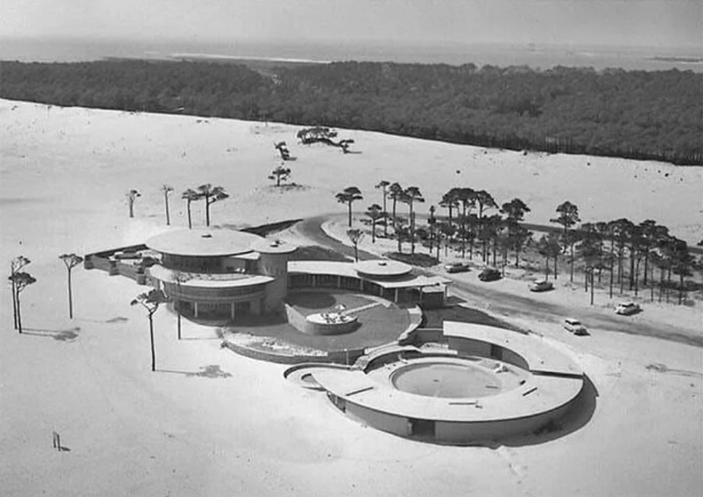 A mid-century modern building with circular design features, surrounded by snow-covered ground, tall trees, and a parking lot with cars.