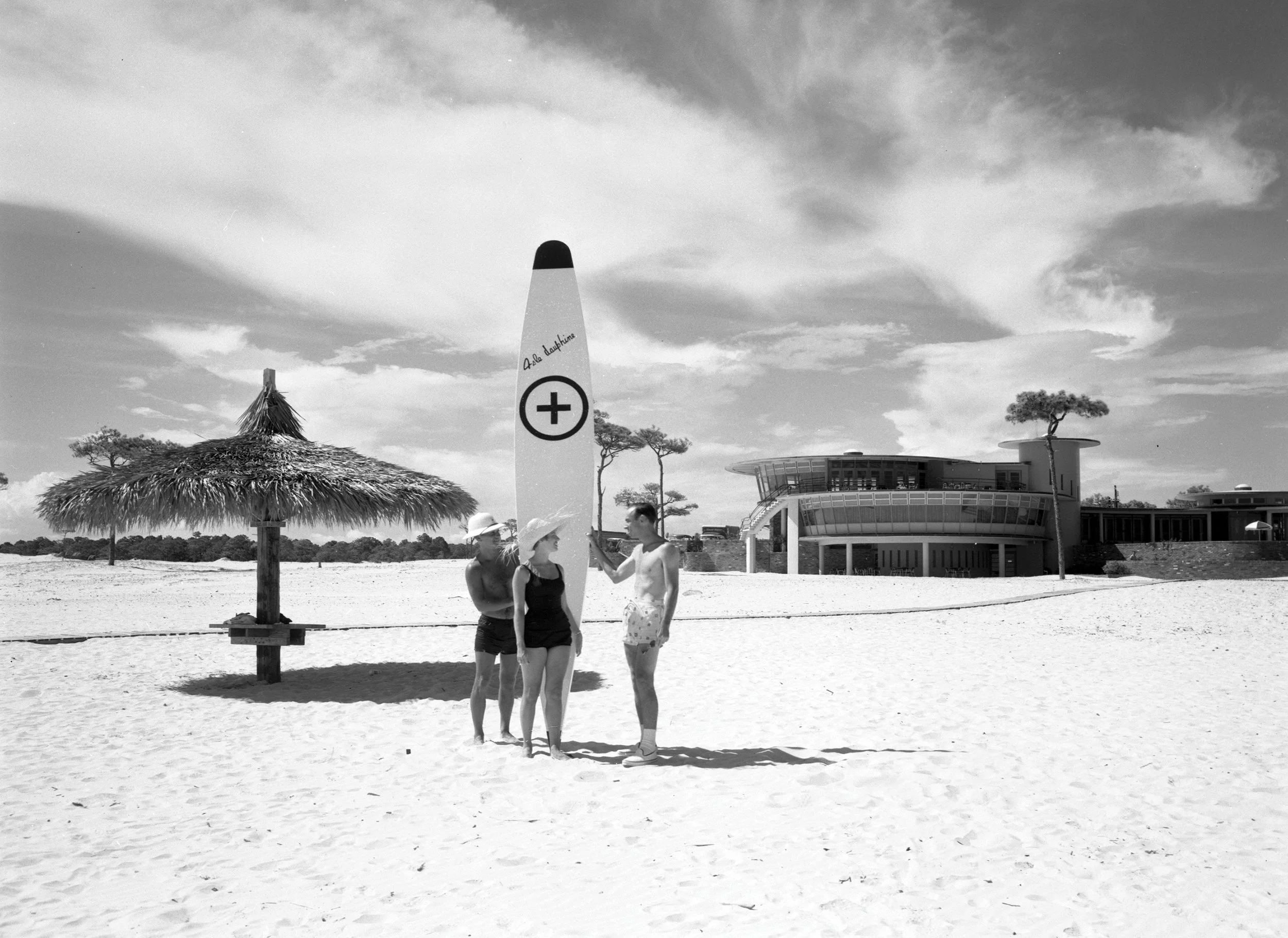 Beach scene in black and white with three people in swimwear, a thatched awning, a sign with a medical cross, trees, and modern architecture building in the background.