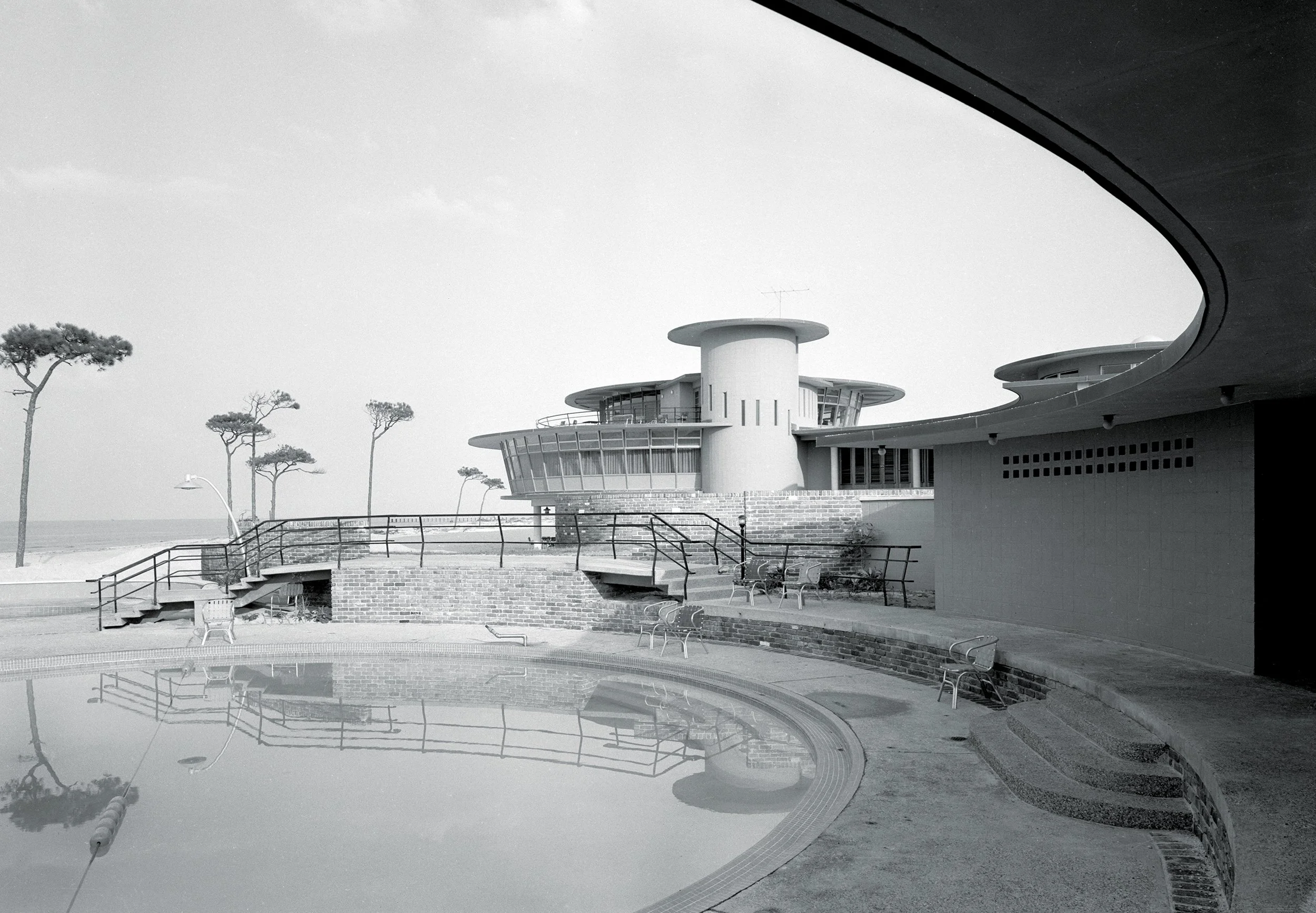 A black and white photo of a modern building with round architectural features, a balcony, and large windows, with a few trees nearby, and a reflection of the building and trees in a swimming pool.
