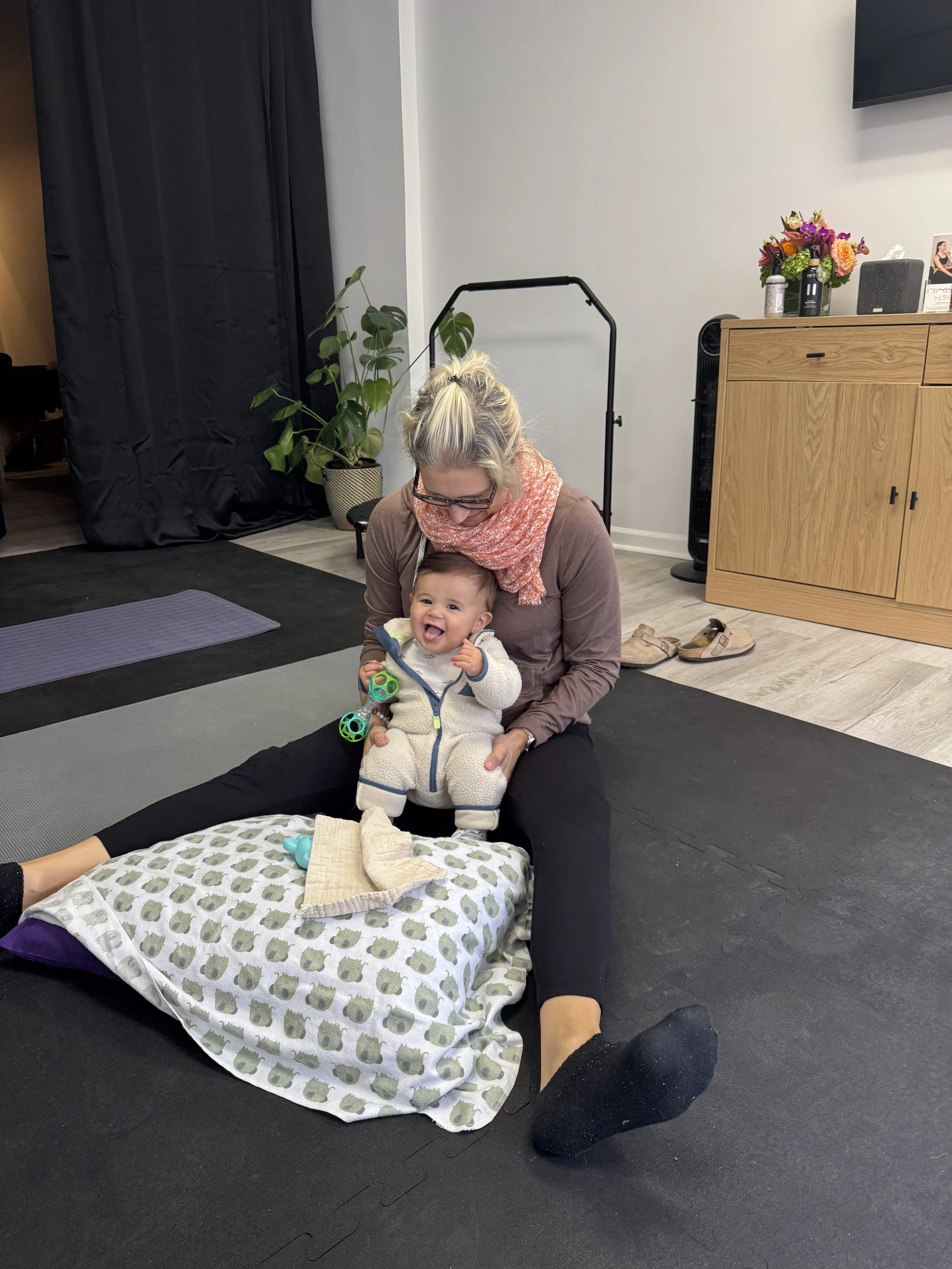 A woman sitting on the floor with her legs apart, holding a smiling baby on her lap. The baby is wearing a white hoodie, and there are baby toys nearby. The room has a wooden cabinet, a tall black fan, an indoor plant, and a black curtain in the background.