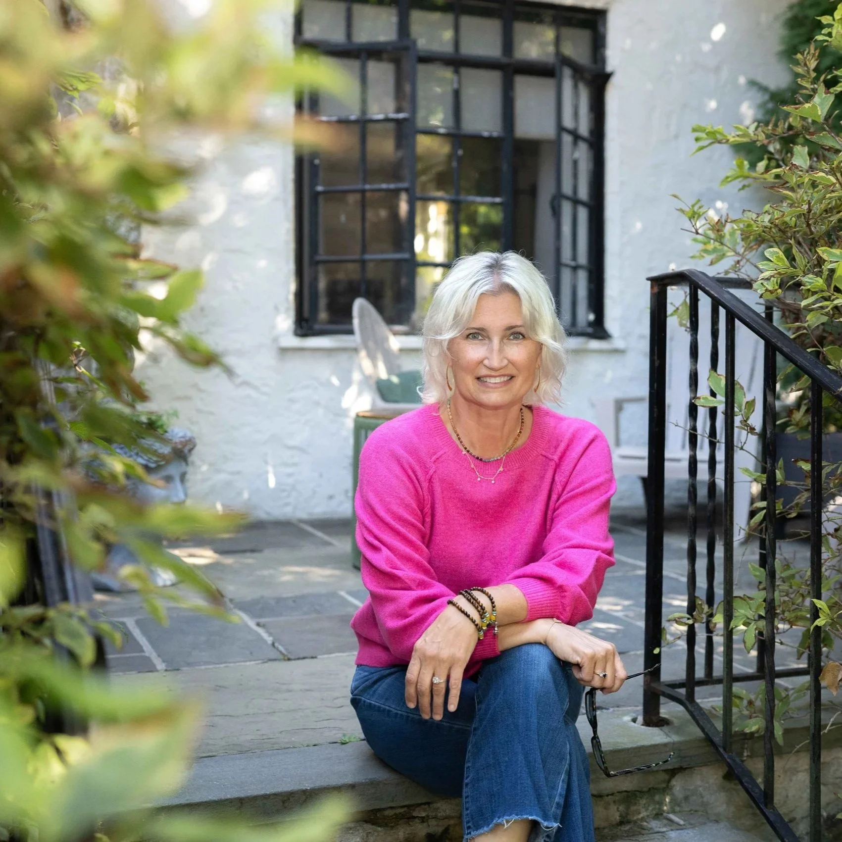 A smiling woman with short blonde hair, wearing a bright pink sweater, sitting on stone steps outside a house with a white stucco wall and black window frames. She's surrounded by greenery and leaning on the black railing.