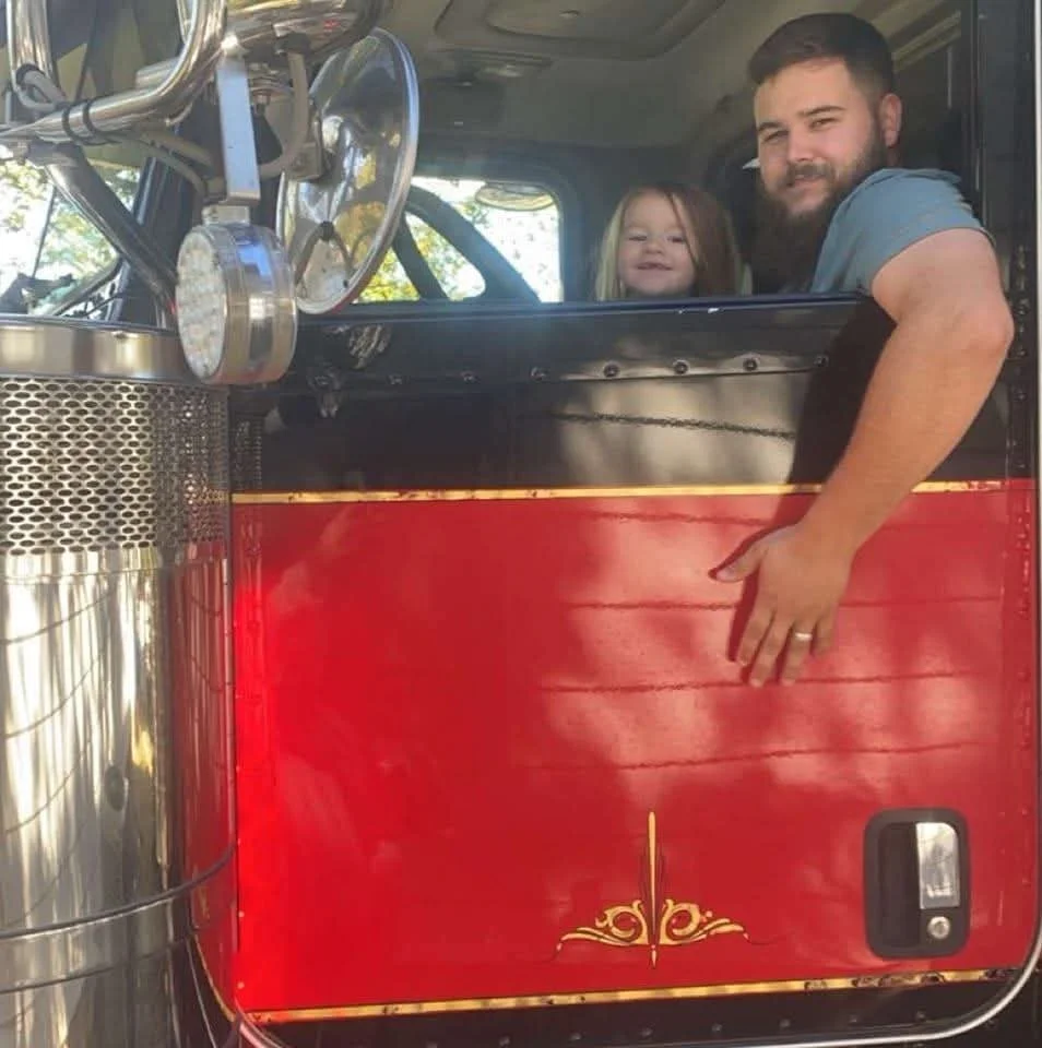A man and a young girl are sitting inside a fire truck, with the man leaning out of the window and the girl smiling behind him.