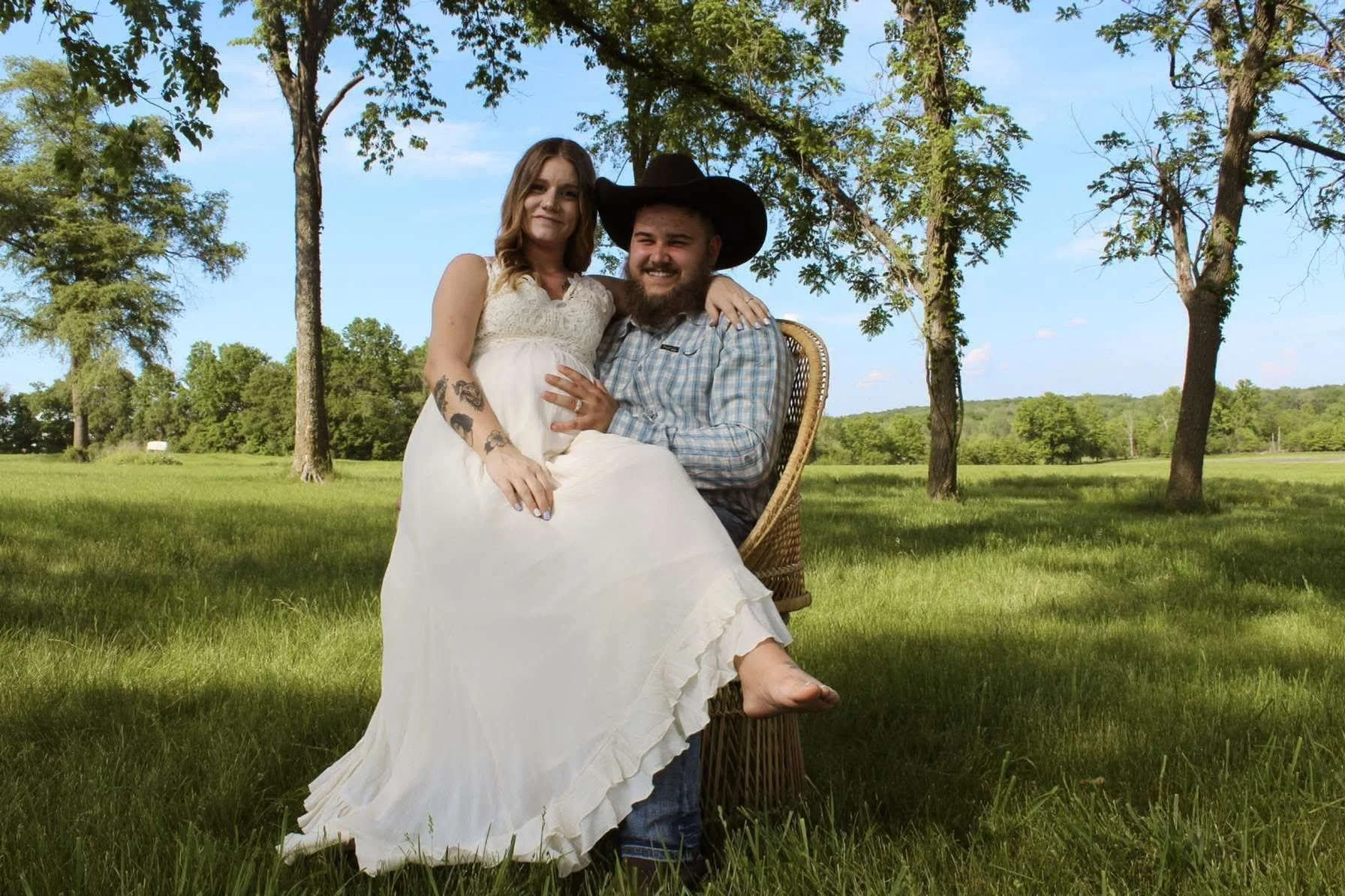 A man in a plaid shirt and cowboy hat sitting on a wicker chair in a grassy field, holding a woman in a white dress who is sitting on his lap with her arm around his neck, surrounded by green trees and a blue sky.