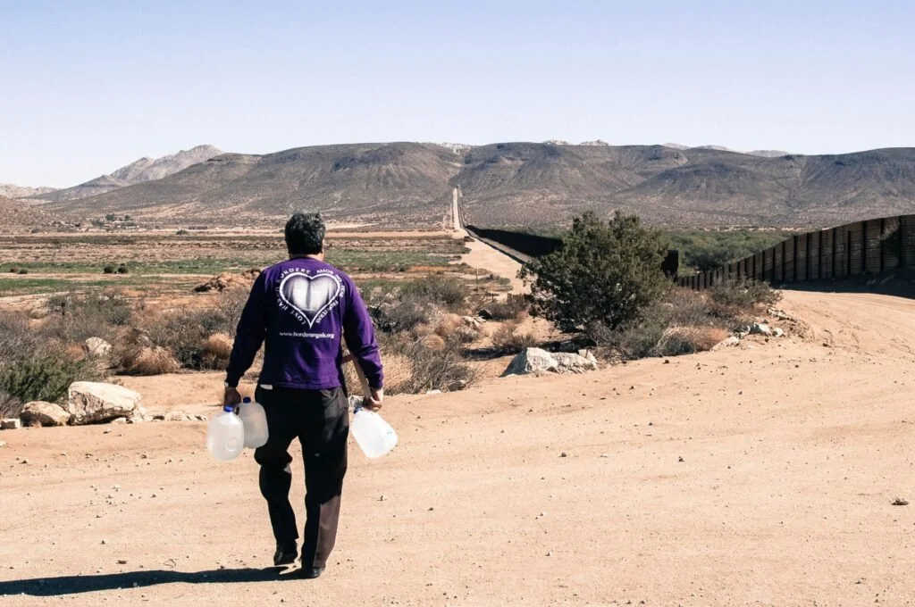 A person walking alone in a desert landscape, carrying large water containers, with mountains and a border fence in the background.