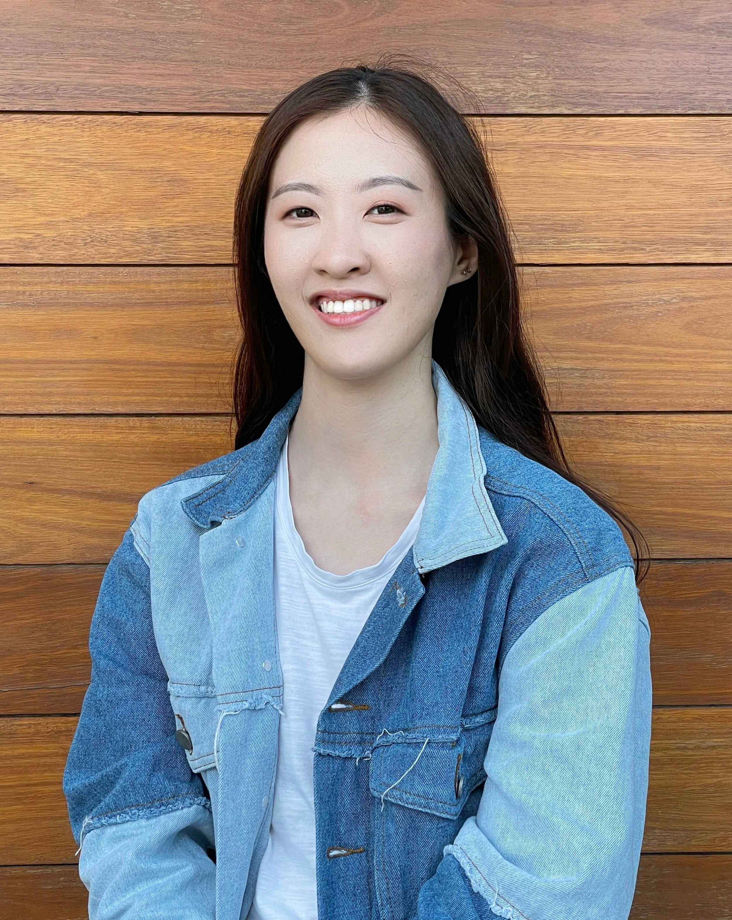 A young woman with long dark hair smiling, wearing a denim jacket over a white shirt, standing in front of a wooden wall.