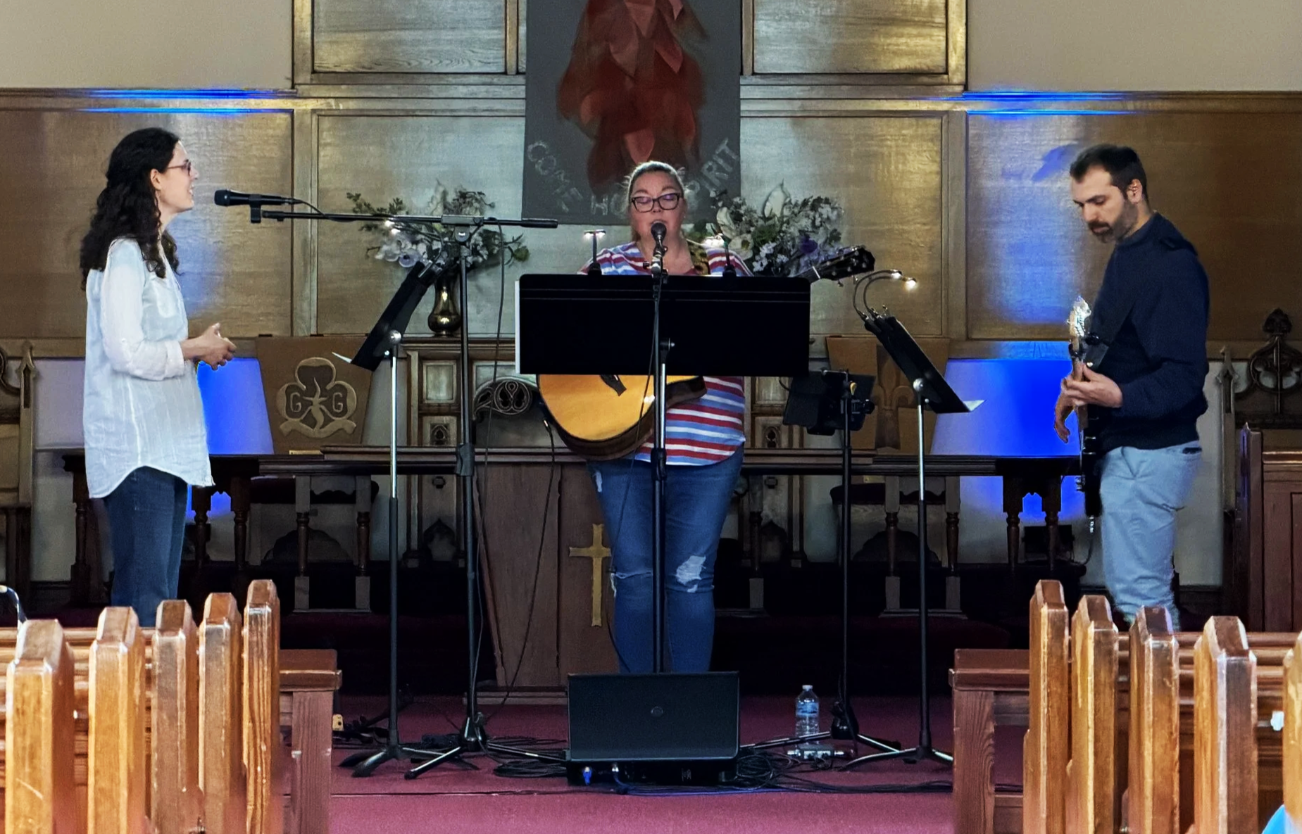 Three people performing music on a church stage with a woman singing and playing guitar, and a man playing bass, while a woman on the left appears to be singing or speaking. The background has a religious-themed painting and floral decorations.