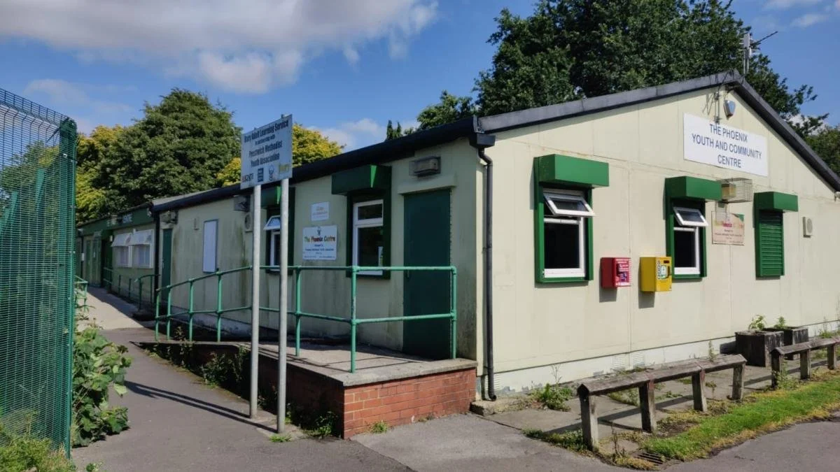 Single-story beige building with green window awnings and signs, labeled 'The Phoenix Youth and Community Centre', with a small ramp in the front and a bench outside, under a partly cloudy sky.