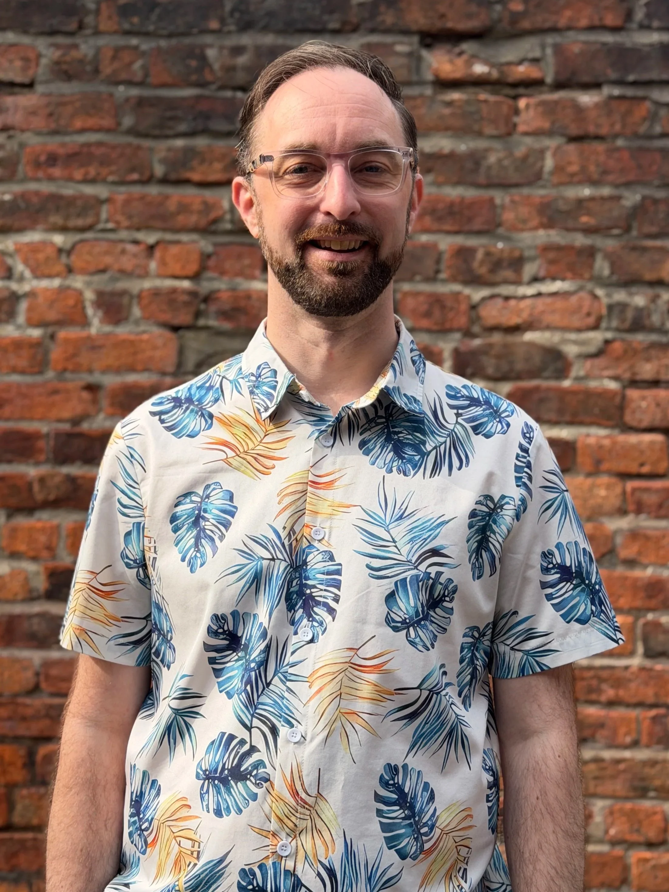 Joe - A smiling man with glasses and a beard, wearing a tropical patterned shirt, stands in front of a brick wall.