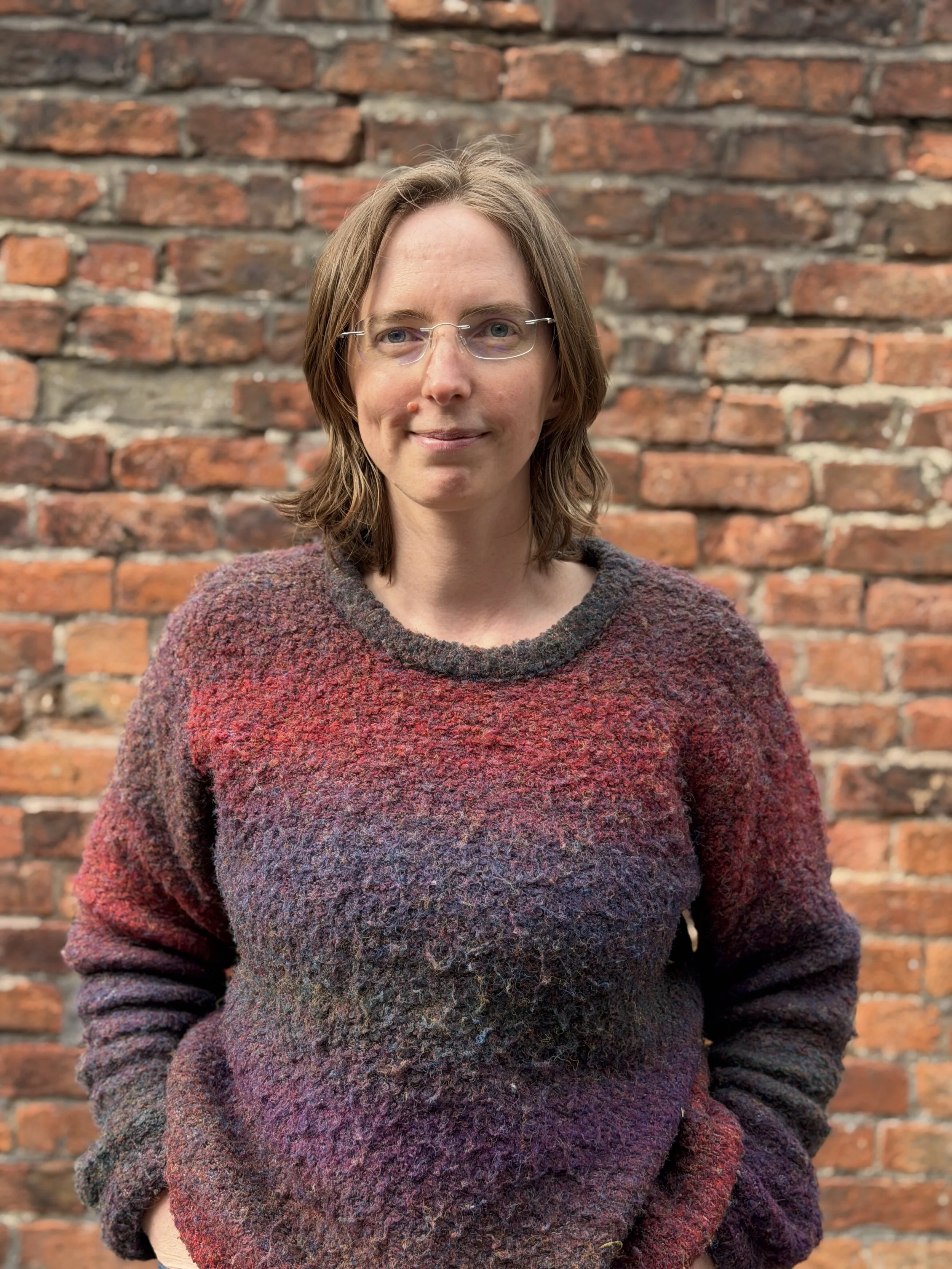 Lianna - Woman with glasses and shoulder-length brown hair standing in front of a brick wall, wearing a multicolored textured sweater.