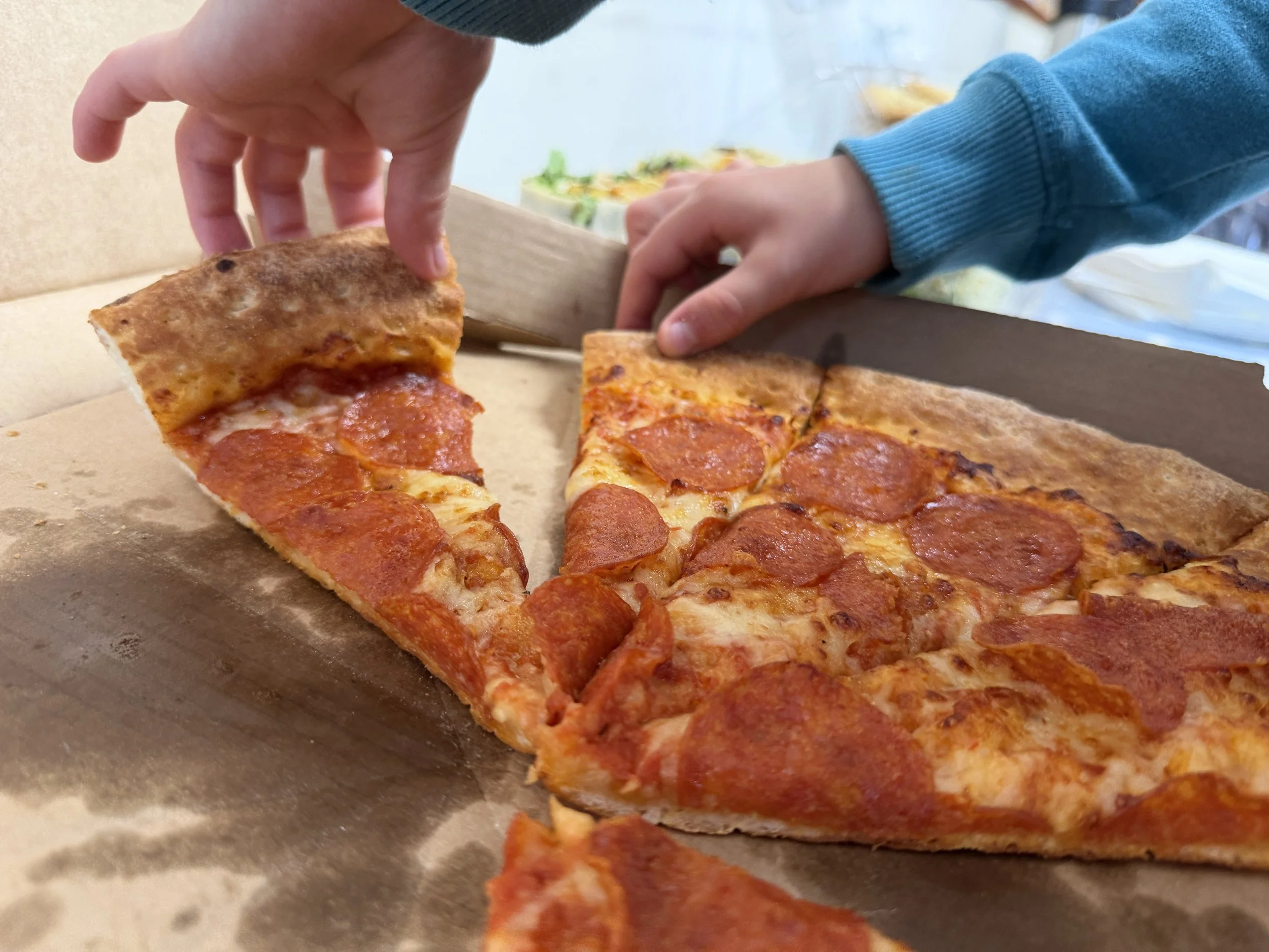 Person in a blue sweatshirt slicing a pepperoni pizza in a cardboard box.