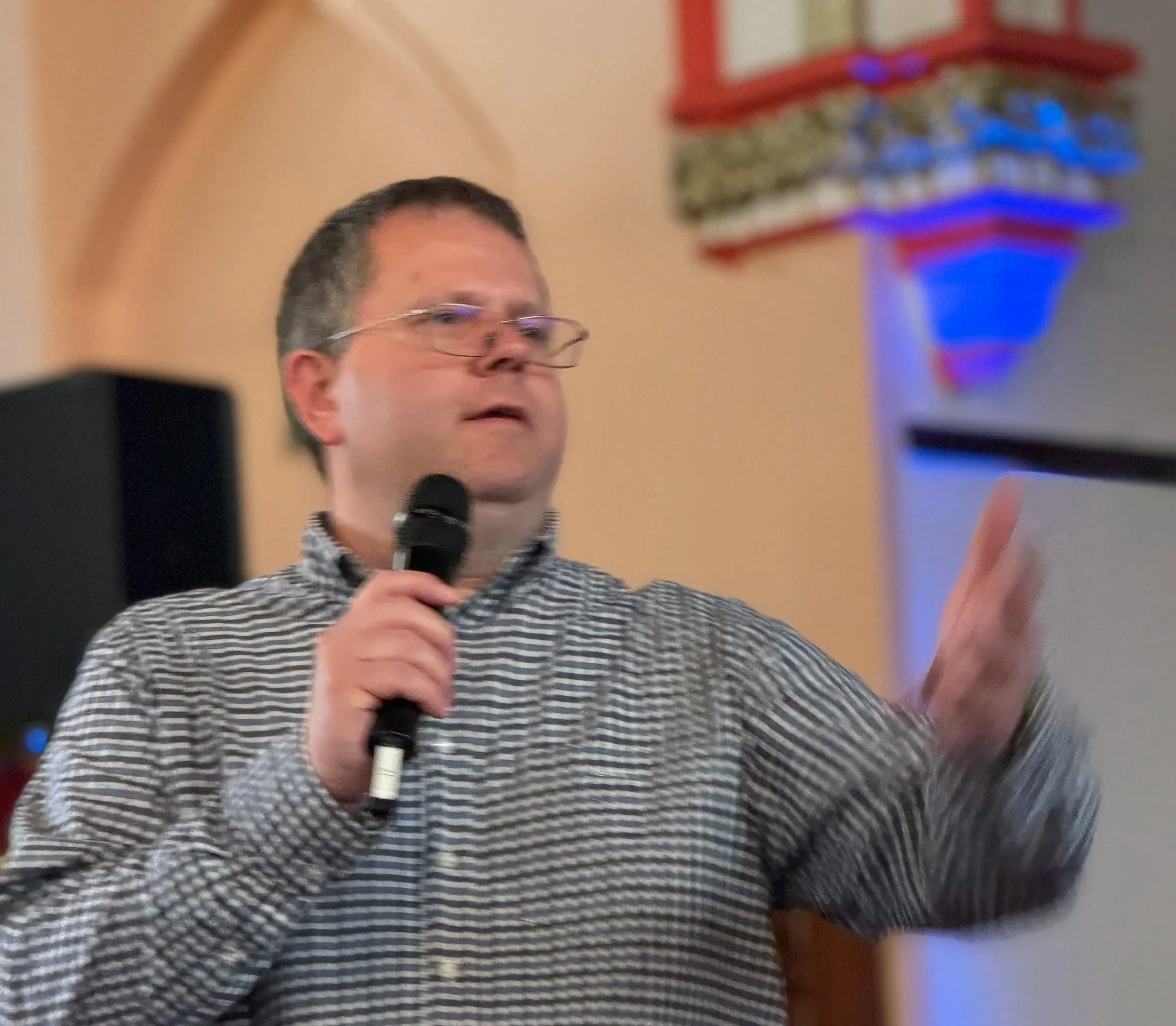 A person holding a microphone and speaking, wearing glasses and a striped shirt, with a blurred background that includes a colorful sign and decorations.