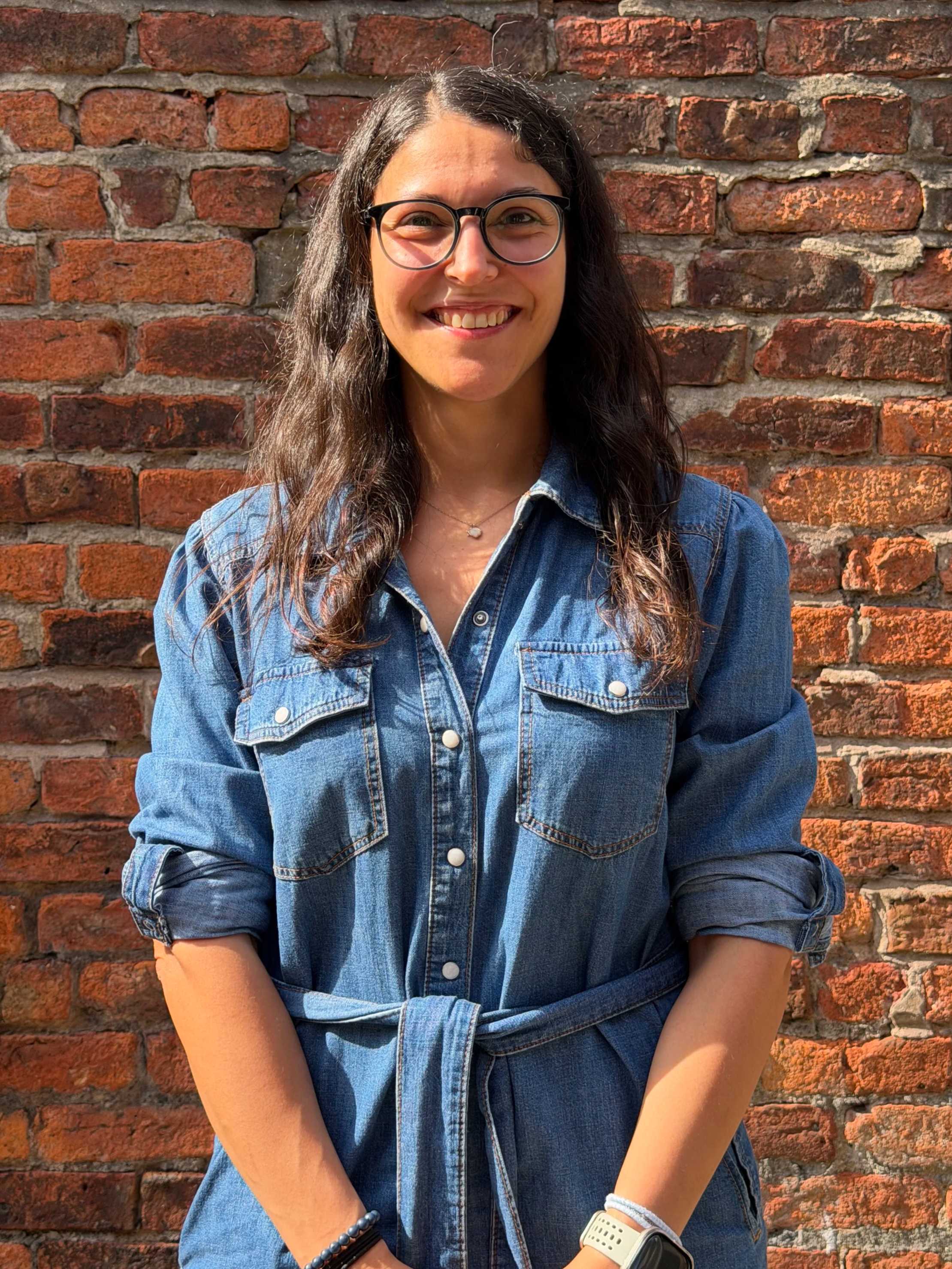 Sandra - A young woman with glasses and long dark hair standing in front of a red brick wall, smiling at the camera, wearing a denim jacket and accessories on her wrists.