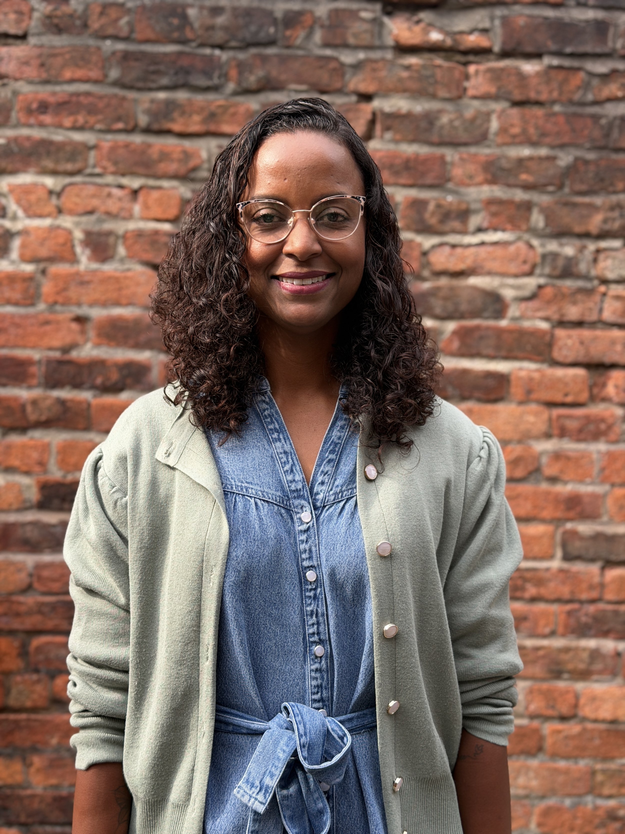 Marcia - A woman with curly hair and glasses standing in front of a red brick wall, wearing a denim shirt with a light green cardigan and a tied denim jacket.