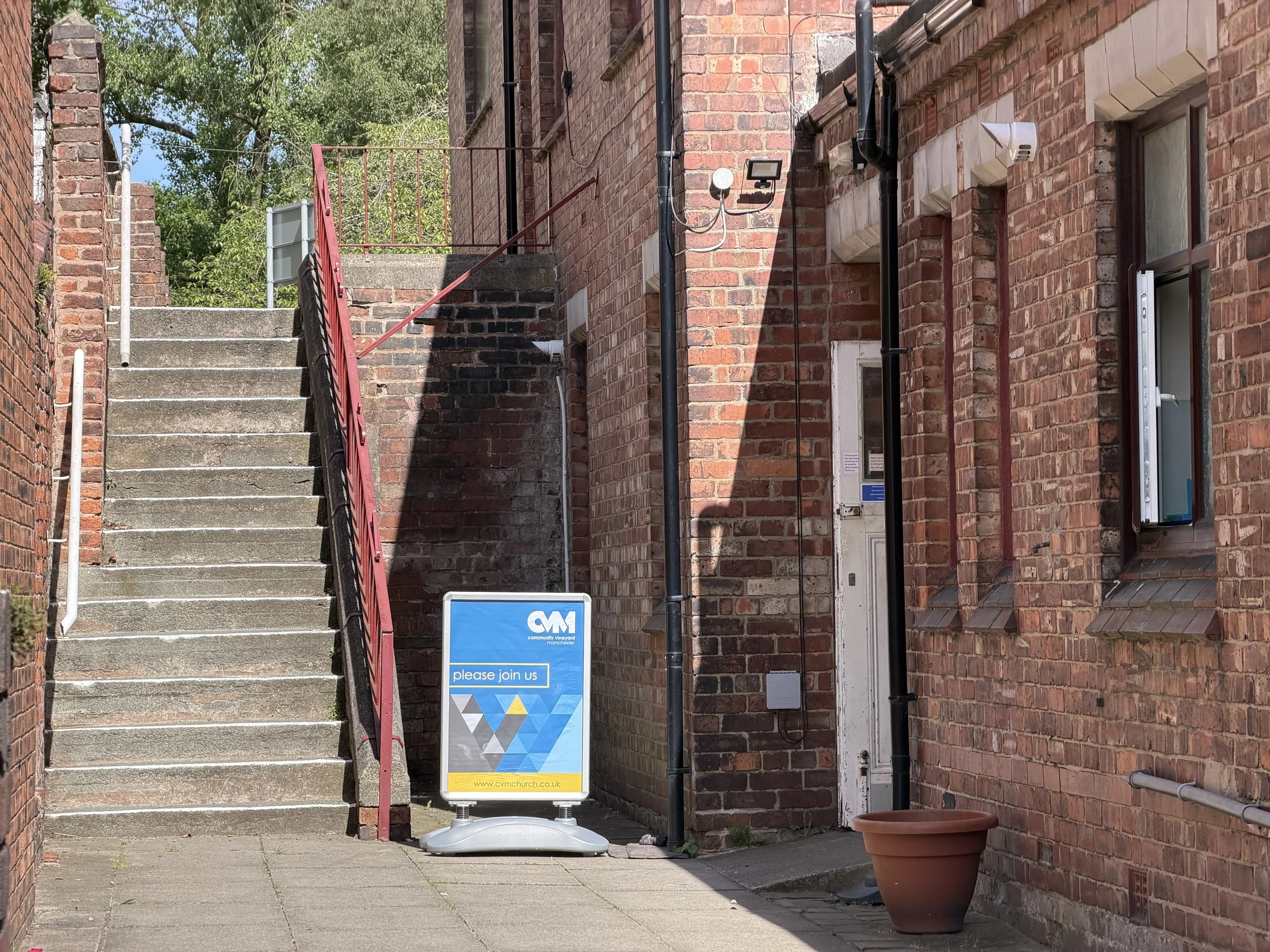 Brick building with stairs leading up, black and red railings, and a sign that reads 'please join us' in front of the building.