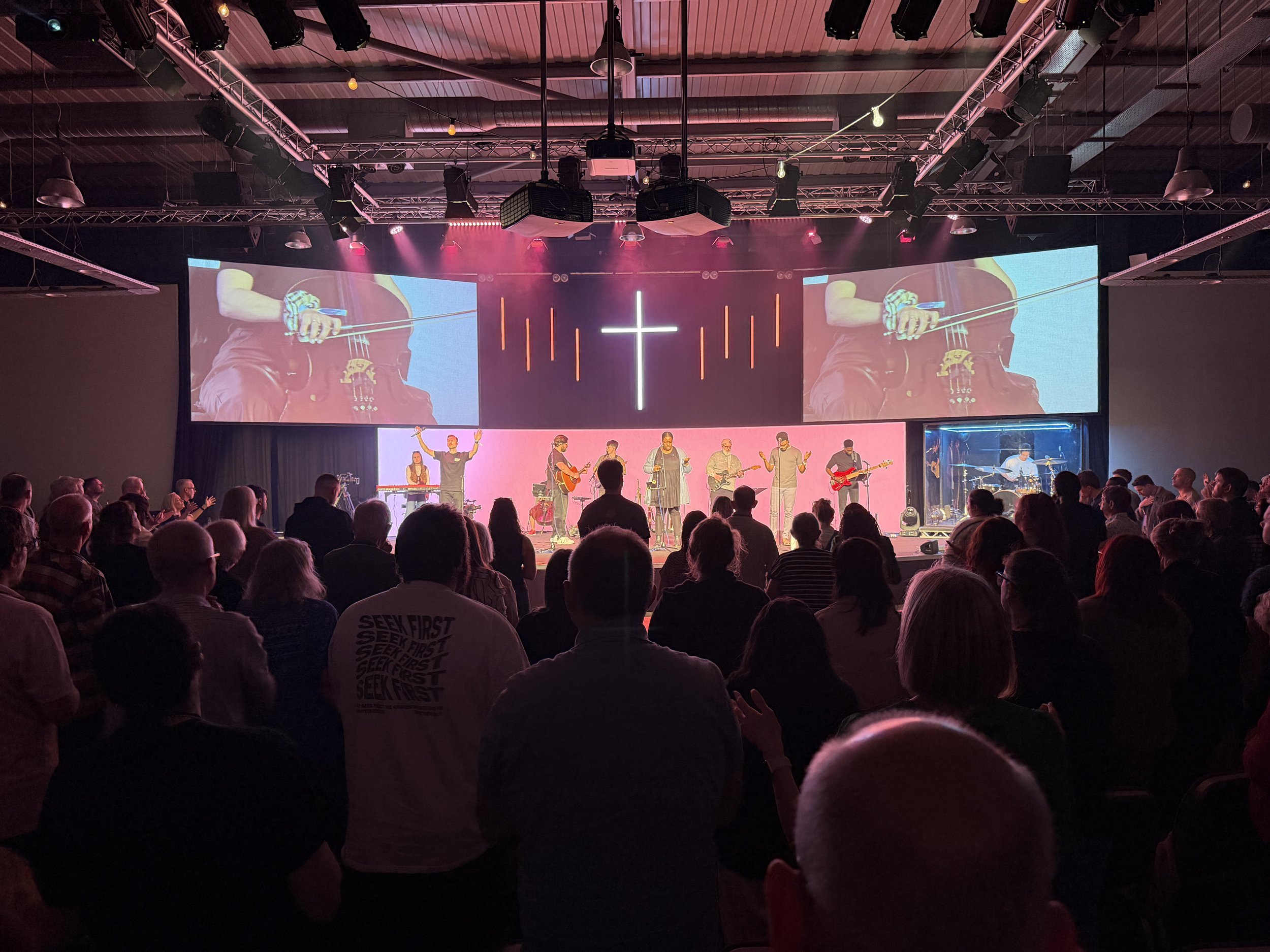 A large indoor concert or event with a stage and a band playing music. The audience is seated facing the stage, and there are large screens displaying a close-up of a musician playing a guitar. The stage has lighting and a cross symbol illuminated at the center.