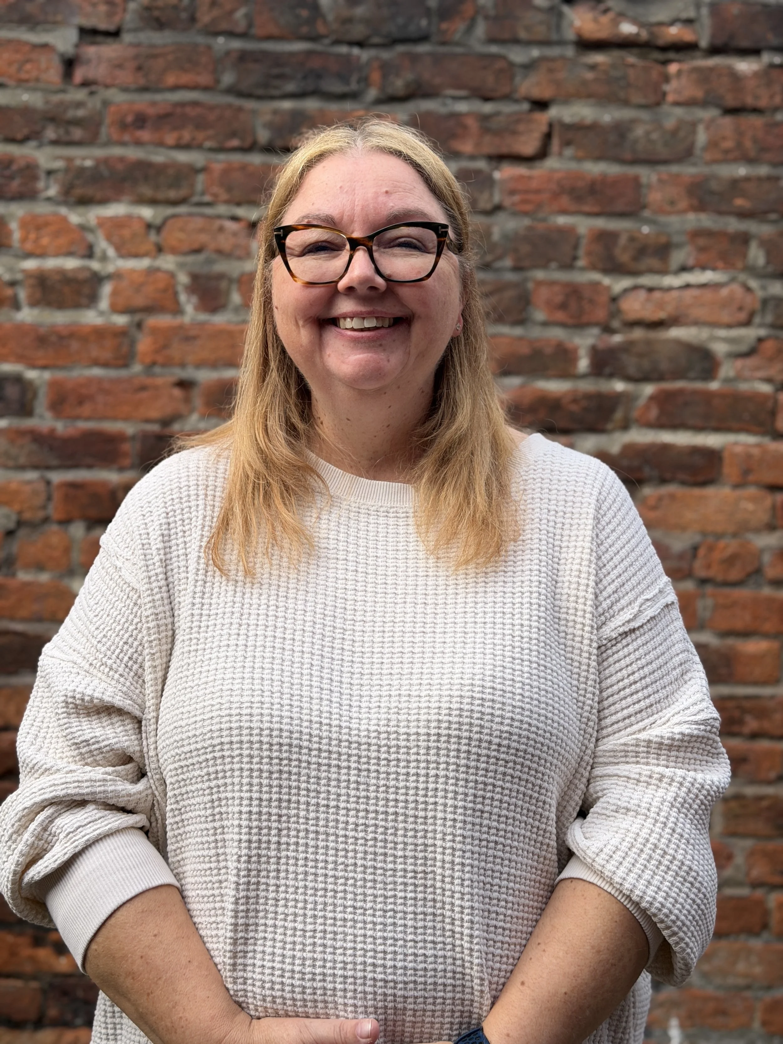 Julie - A woman with glasses smiling, wearing a light-colored sweater, standing in front of a red brick wall.