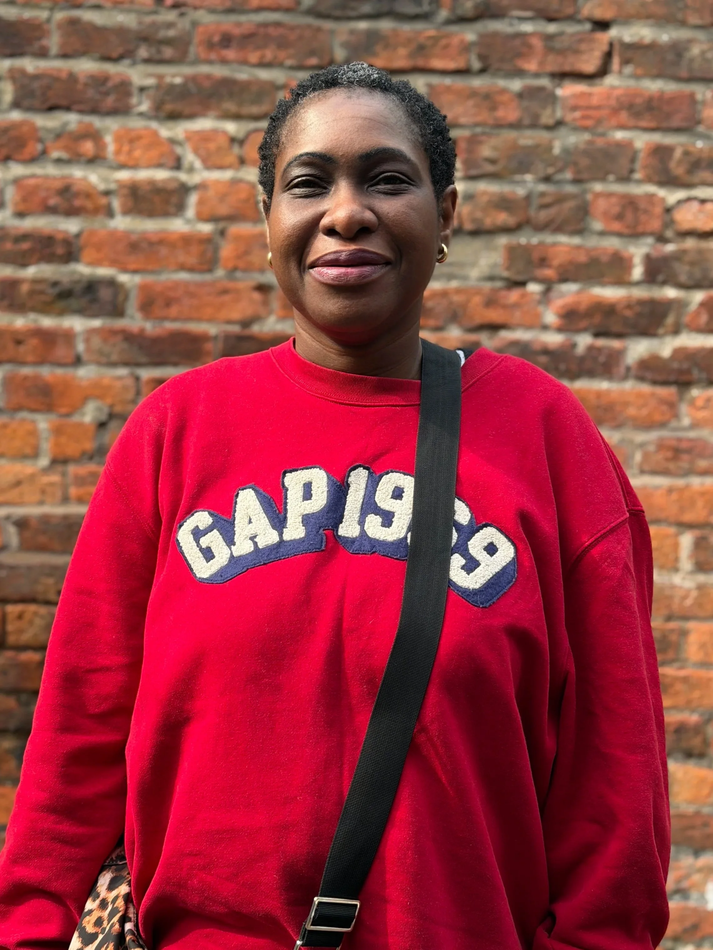 Kes - A woman with short curly hair smiling in front of a brick wall, wearing a red GAP sweatshirt and gold hoop earrings.