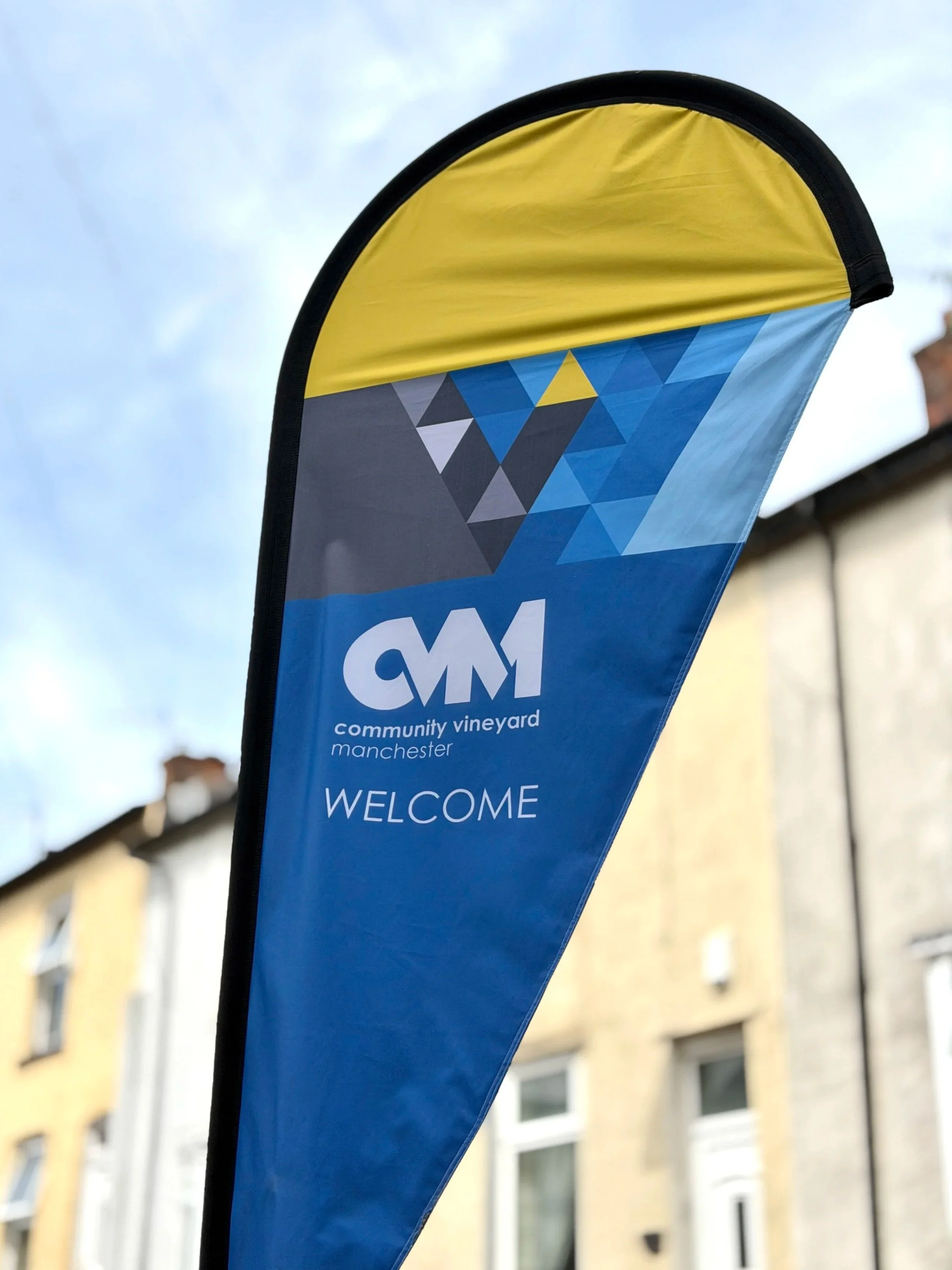 A blue and yellow feather-shaped banner with the logo of Community Vineyard Manchester, featuring geometric shapes and the word 'WELCOME' at the bottom.