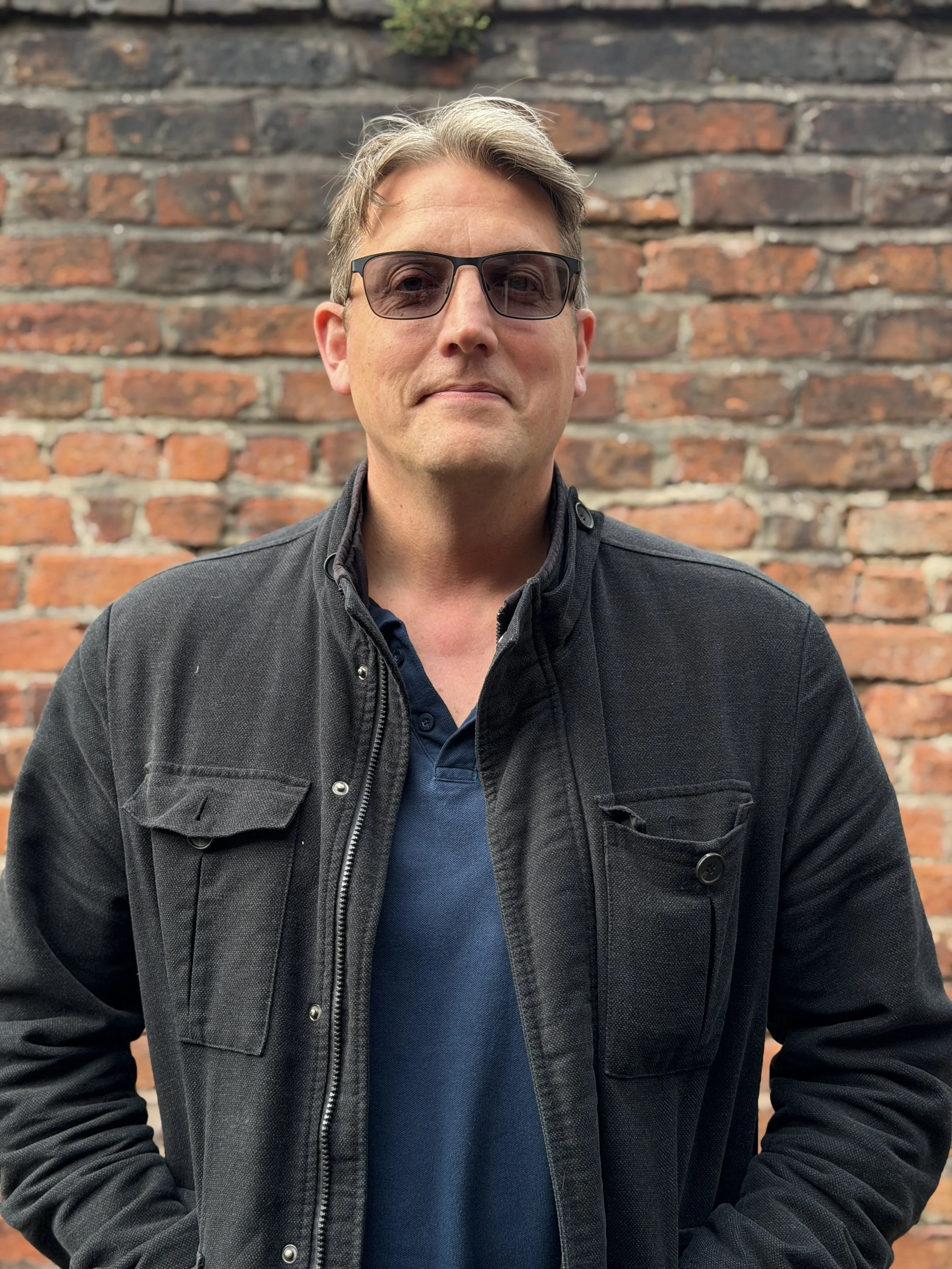 Stefan - A man with smart hair wearing sunglasses, a navy blue shirt, and a black jacket standing in front of a brick wall.