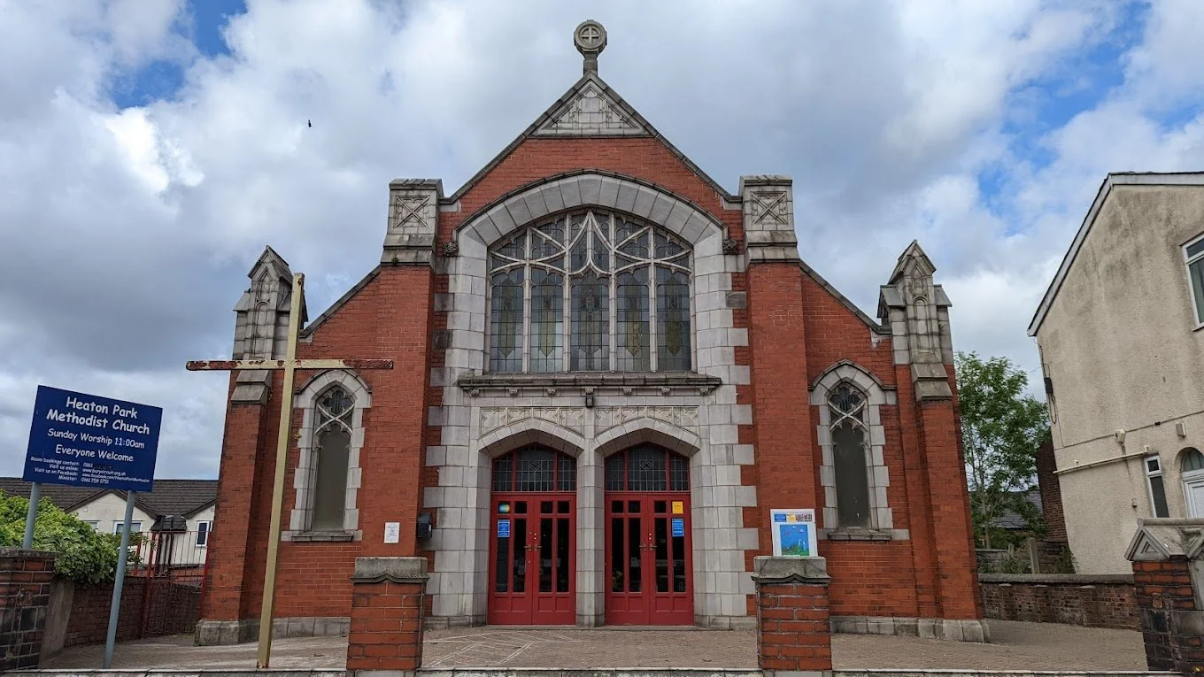 Front view of a red brick church with large arched stained glass windows and a pointed roof, flanked by smaller gothic windows, with a cross-shaped wooden sculpture and a congregation sign outside.