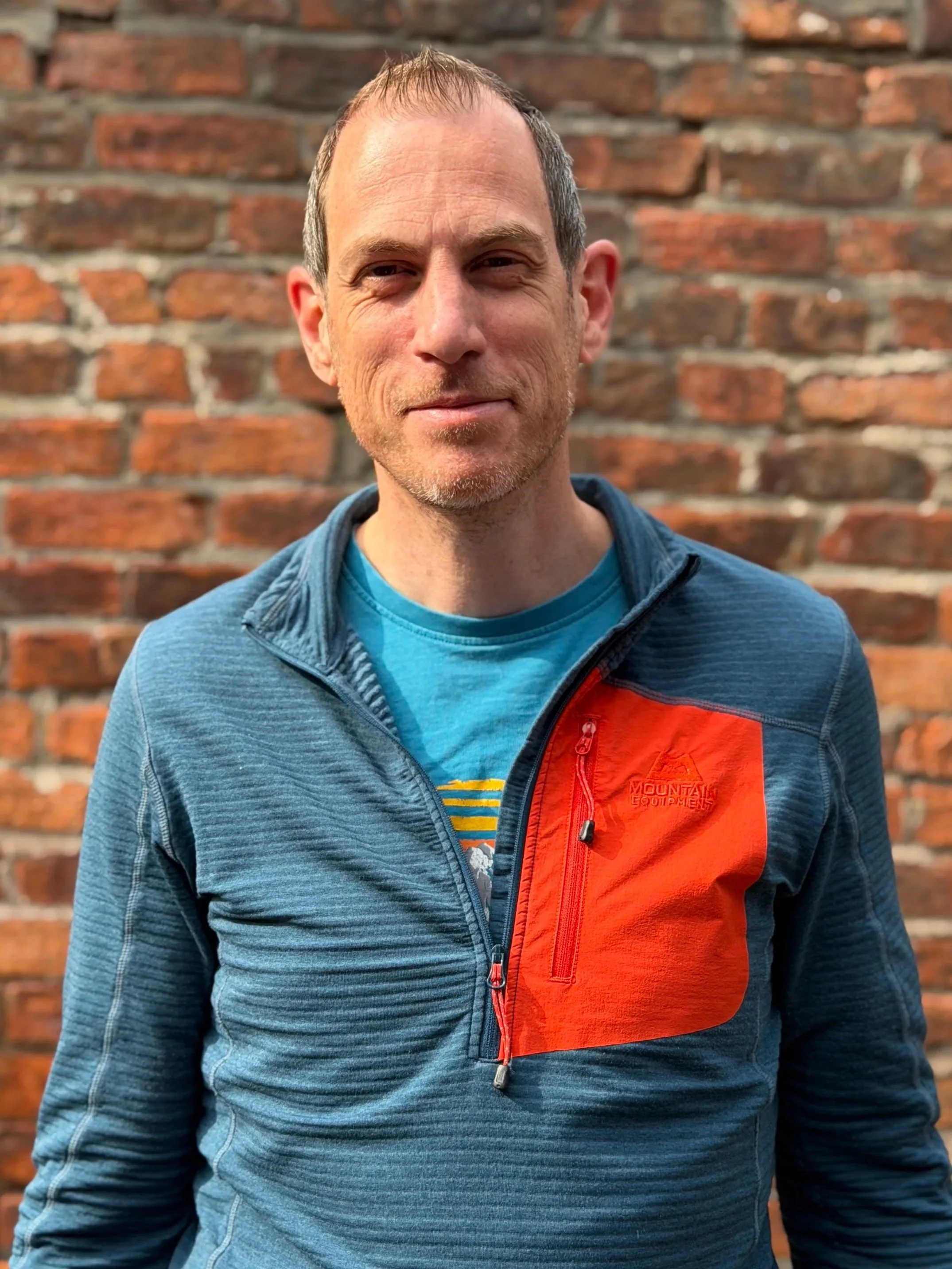 Paul - A man wearing a blue and orange Mountain Equipment jacket standing in front of a red brick wall.