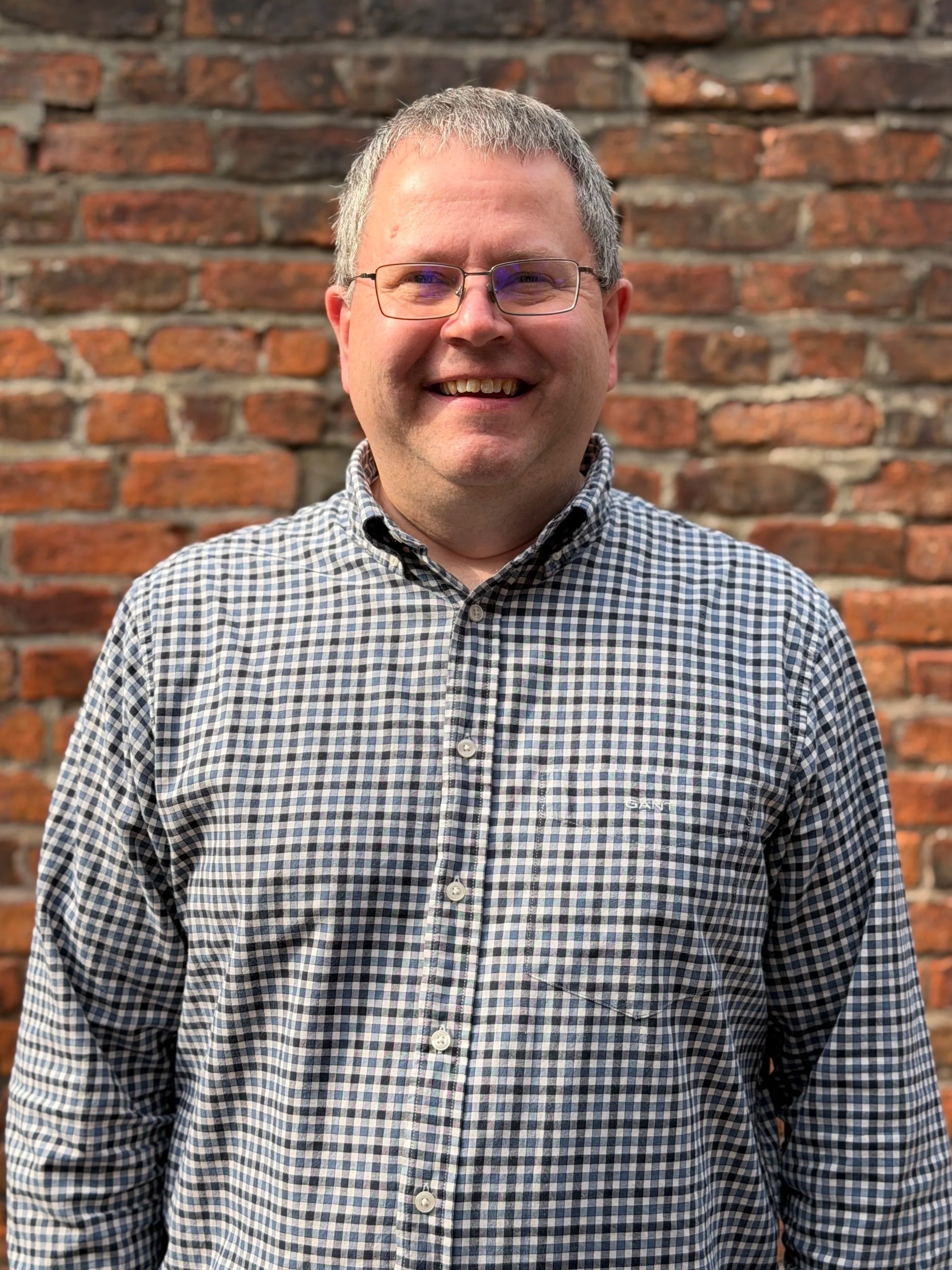 Ross - A man with glasses and short hair smiling, wearing a checkered shirt, standing in front of a brick wall.