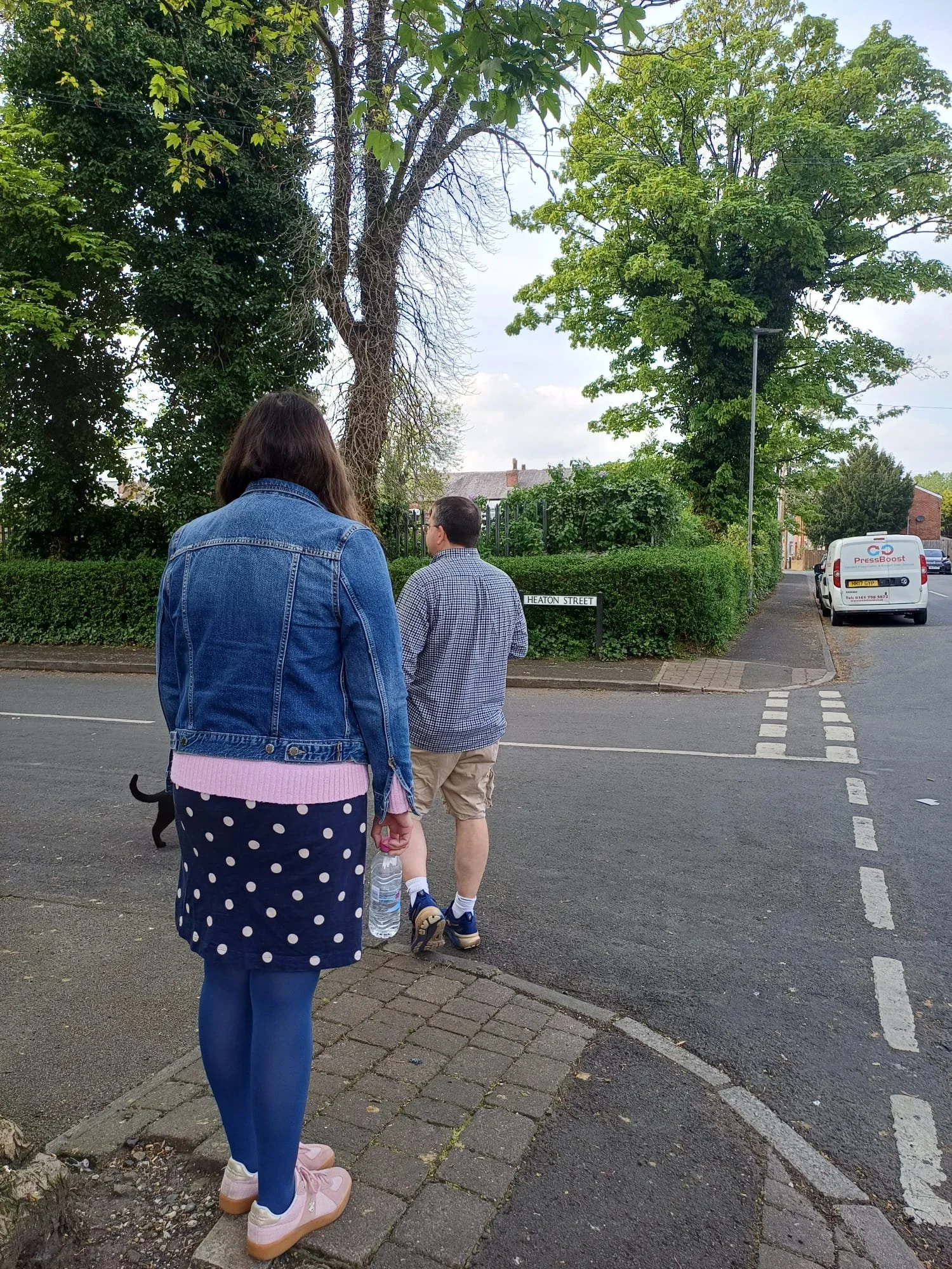 Three people walking on a sidewalk near a park or green space with trees and a street sign that reads 'Heaton Street'. One person is in front, wearing a denim jacket, blue tights, and pink shoes, holding a water bottle. The other two are walking ahead, one in shorts and a plaid shirt, and the other has long hair. A white van is parked along the street.