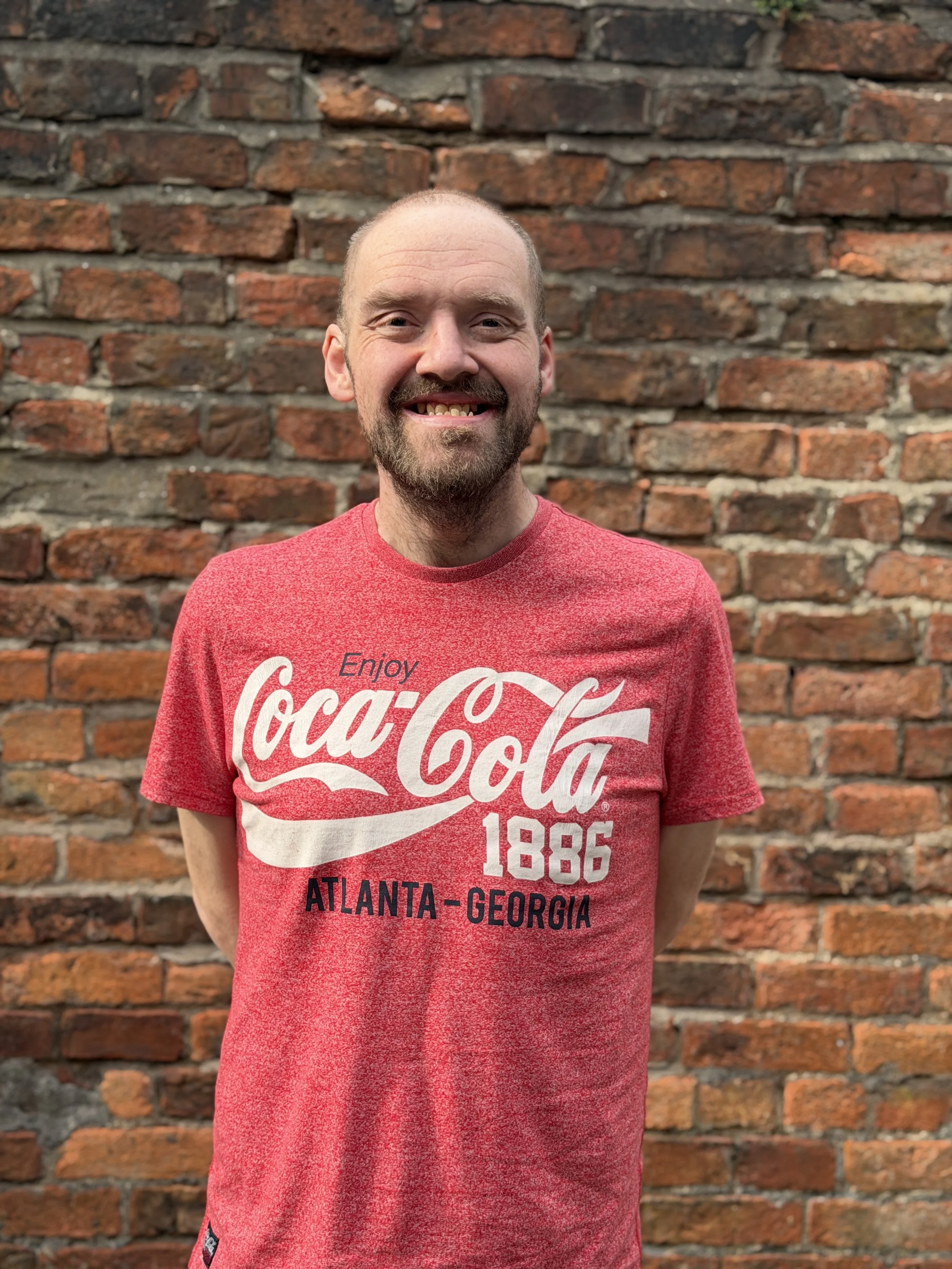 Phil - A smiling man with a beard and short hair standing in front of a brick wall, wearing a red Coca-Cola T-shirt.
