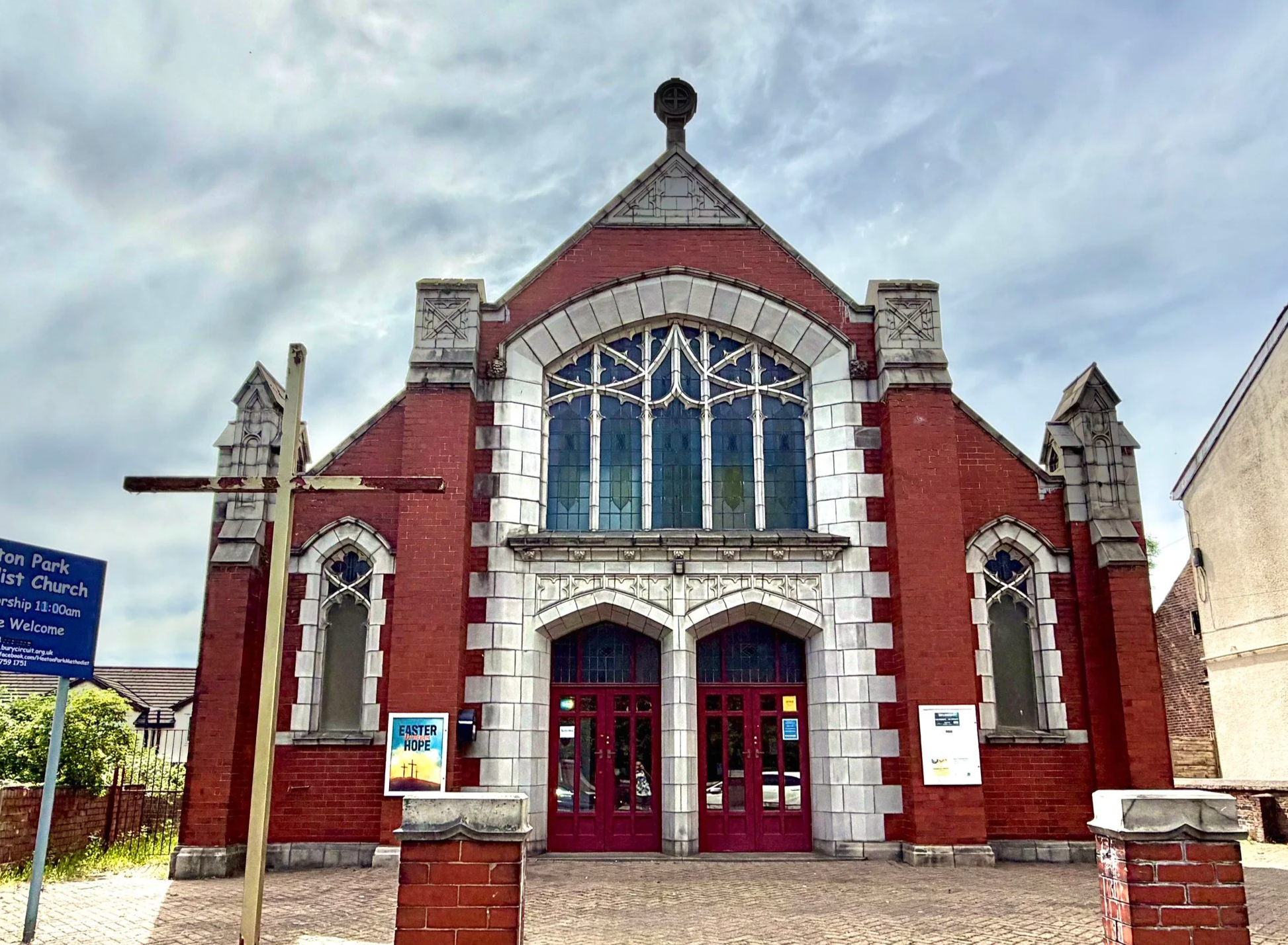 A red brick church with stone accents and stained glass windows, topped with a cross, under a cloudy sky. There's a poster and informational signs near the entrance.