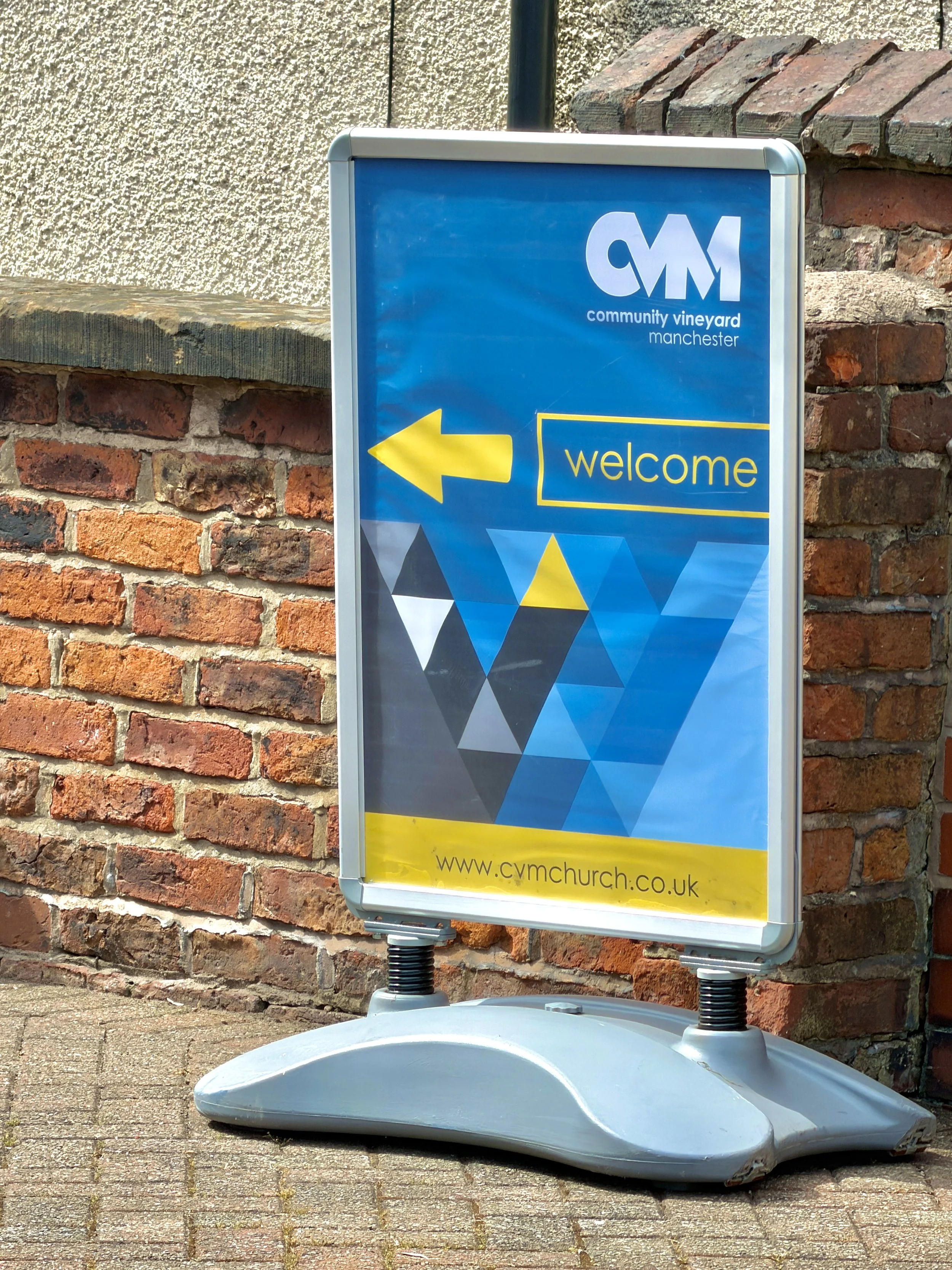 Signboard for Community Vineyard Manchester with a yellow left arrow and the word 'welcome' on a blue background.