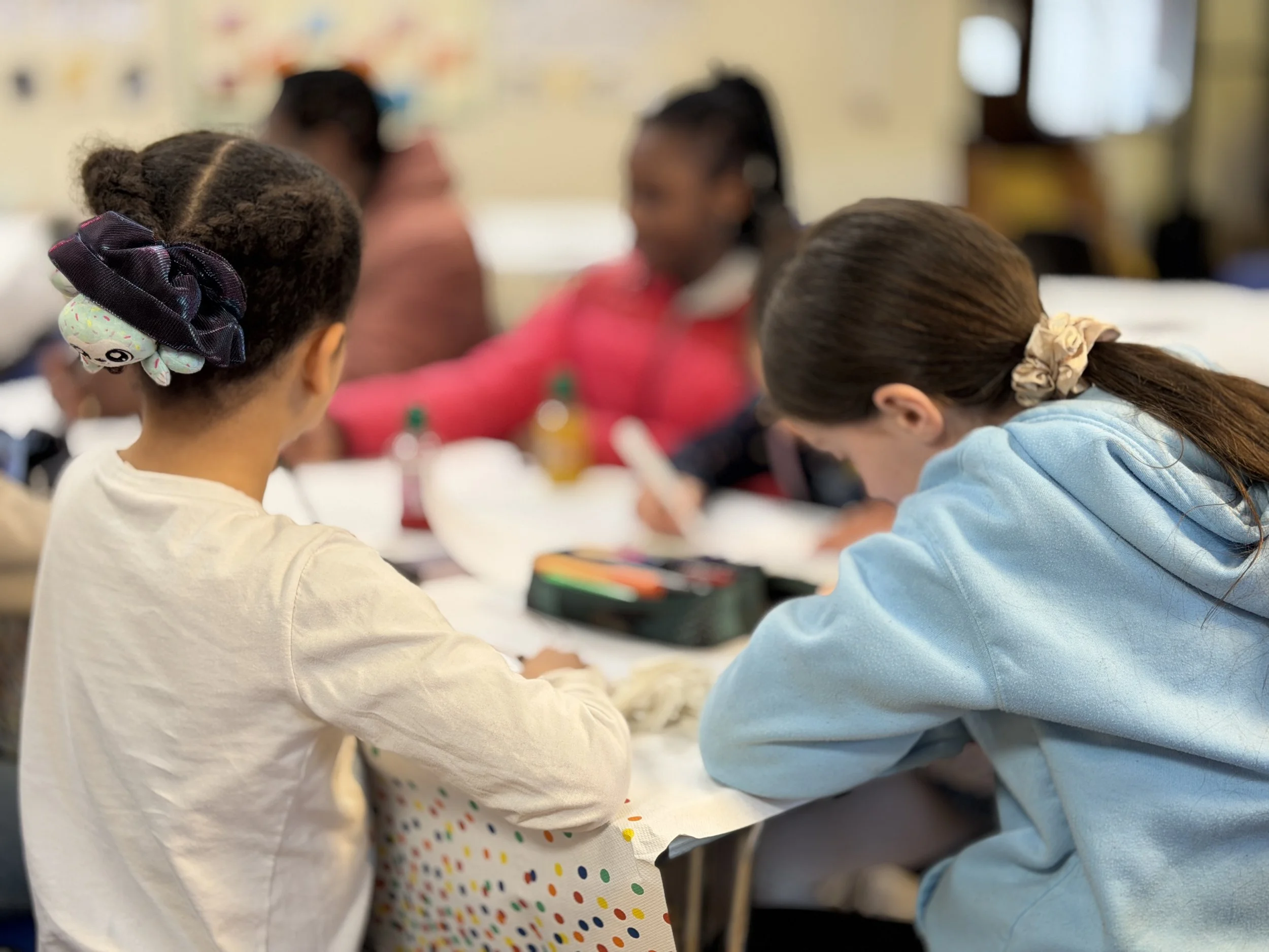 Children sitting at a table in a classroom, focusing on activities, with colorful decorations on the wall in the background.