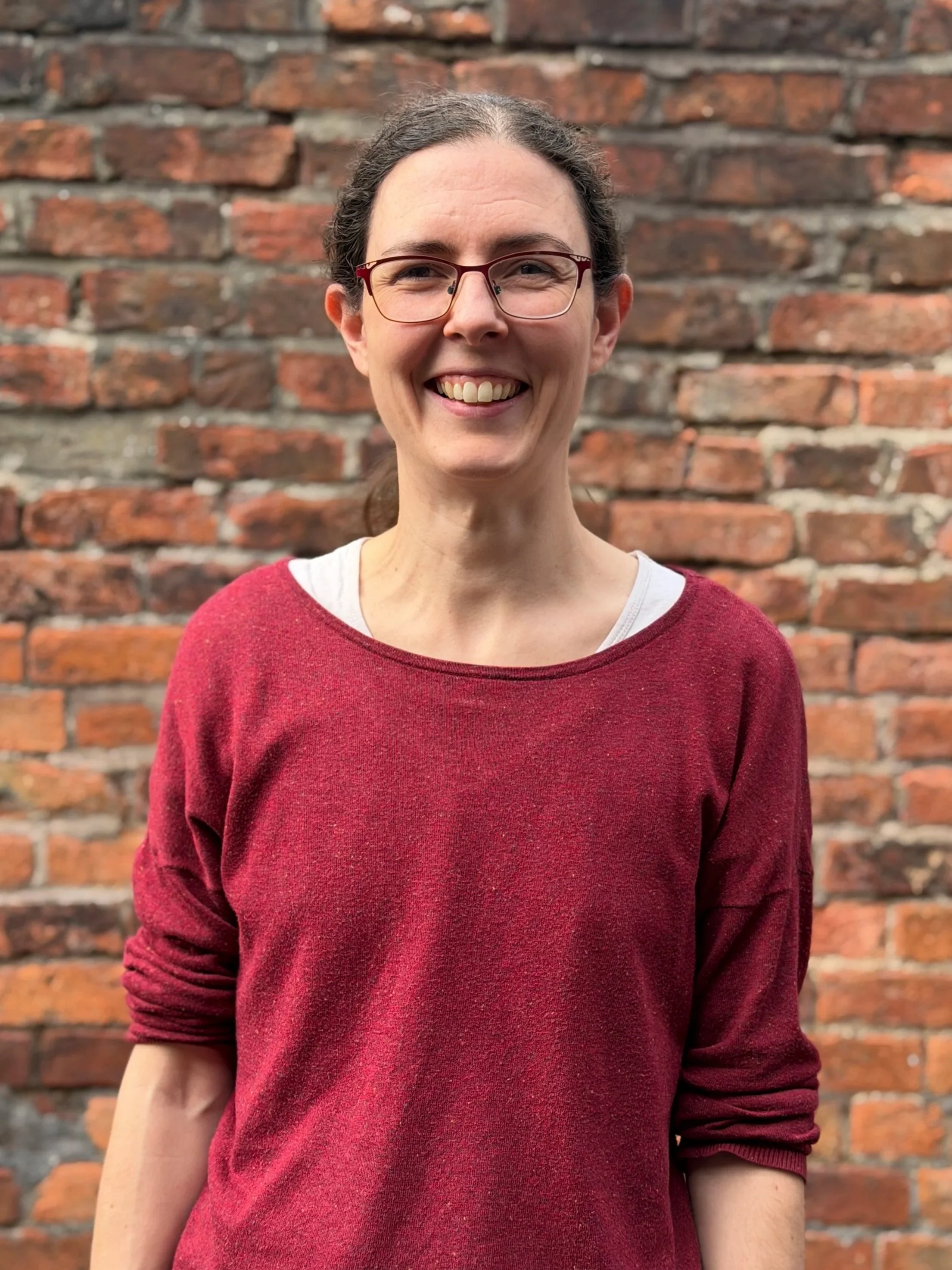 Solenne - Smiling woman with glasses and brown hair tied back, wearing a red sweater over a white shirt, standing in front of a brick wall.