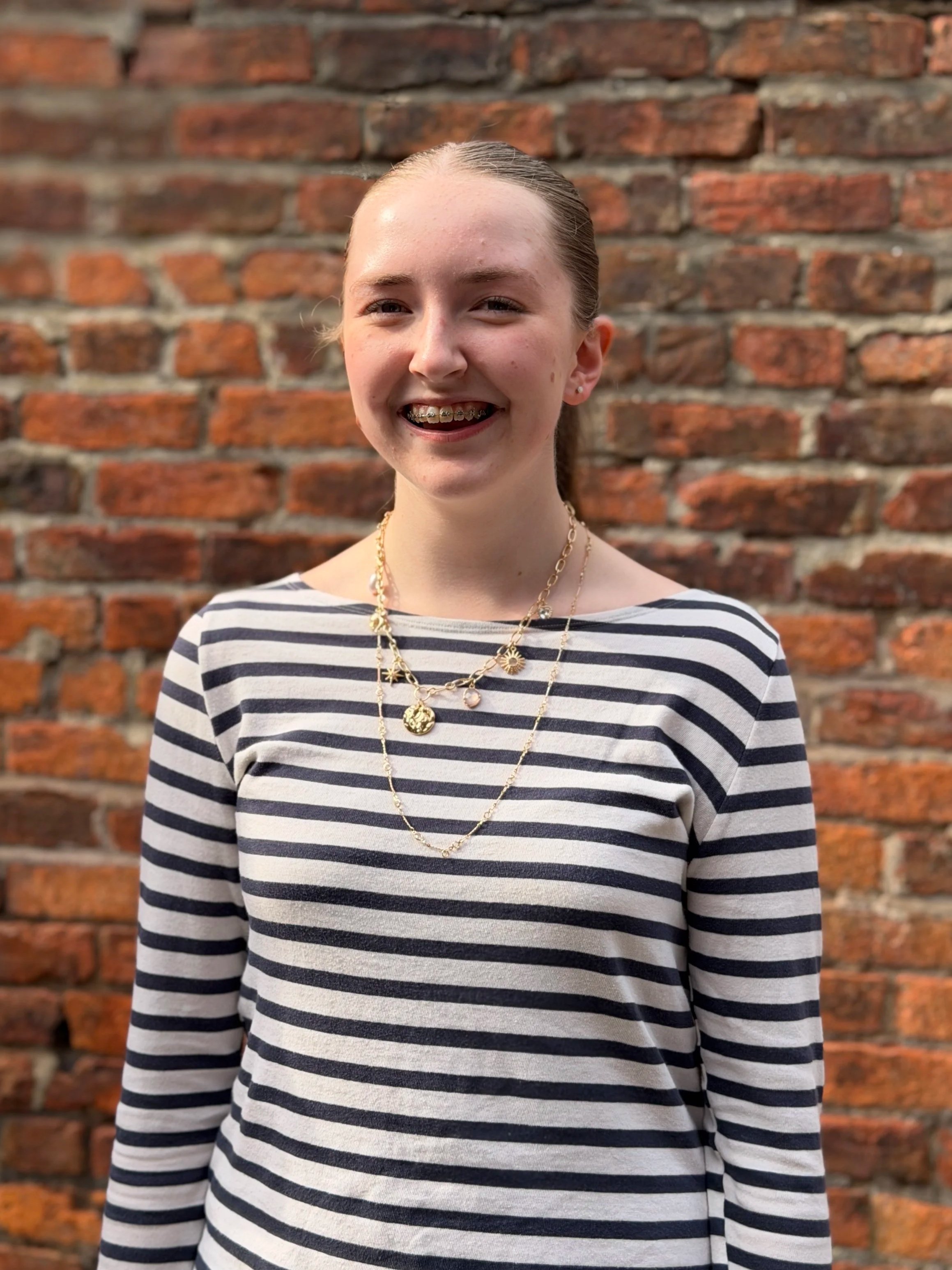 Emily - A young woman with long brown hair tied back, smiling, standing in front of a rustic red brick wall. She is wearing a striped long-sleeve shirt and layered gold necklaces.