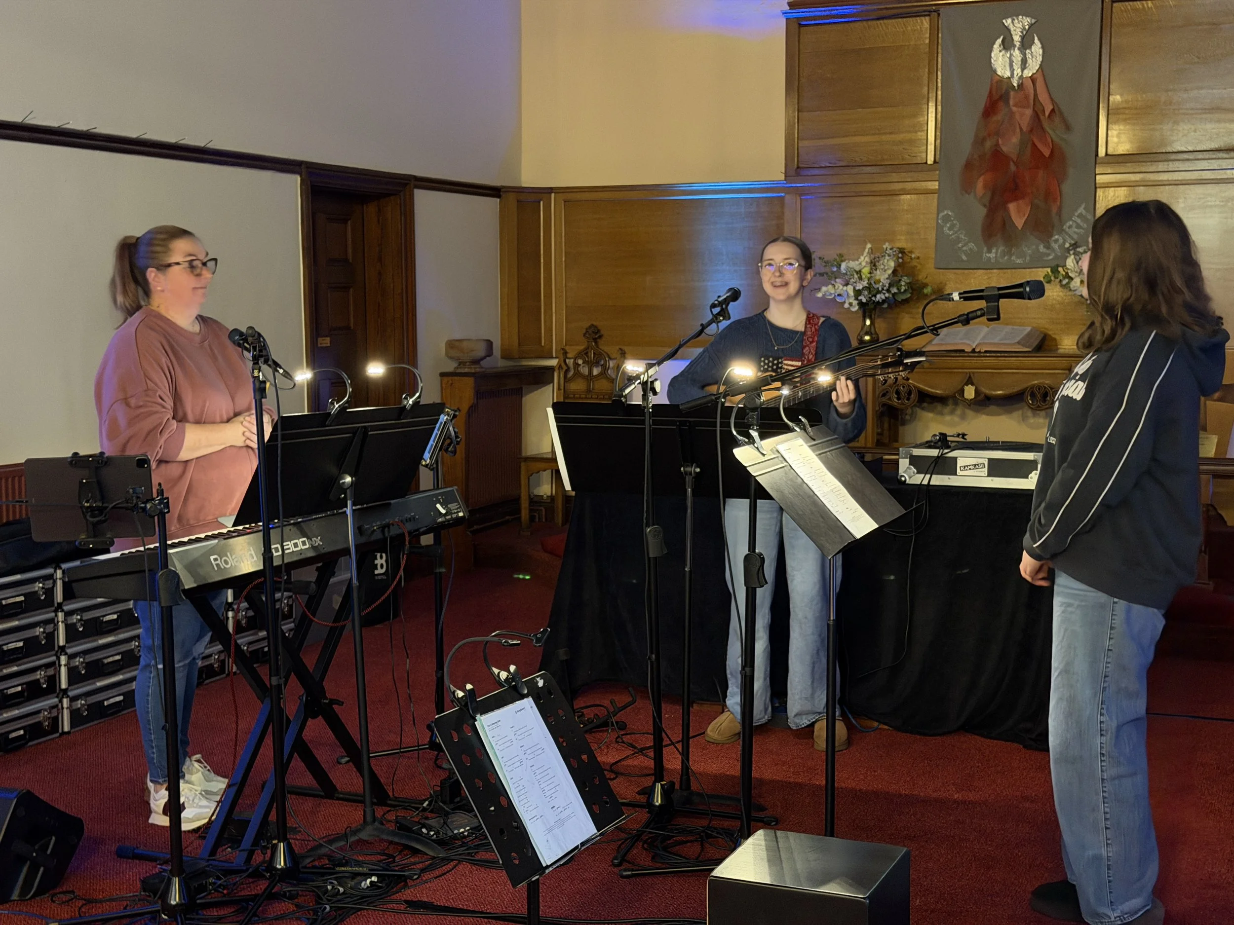 Three women are standing in a church, singing and playing musical instruments, with music stands and microphones in front of them. The church interior features wooden wall panels, a banner with an eagle and a red flower, and floral arrangements.