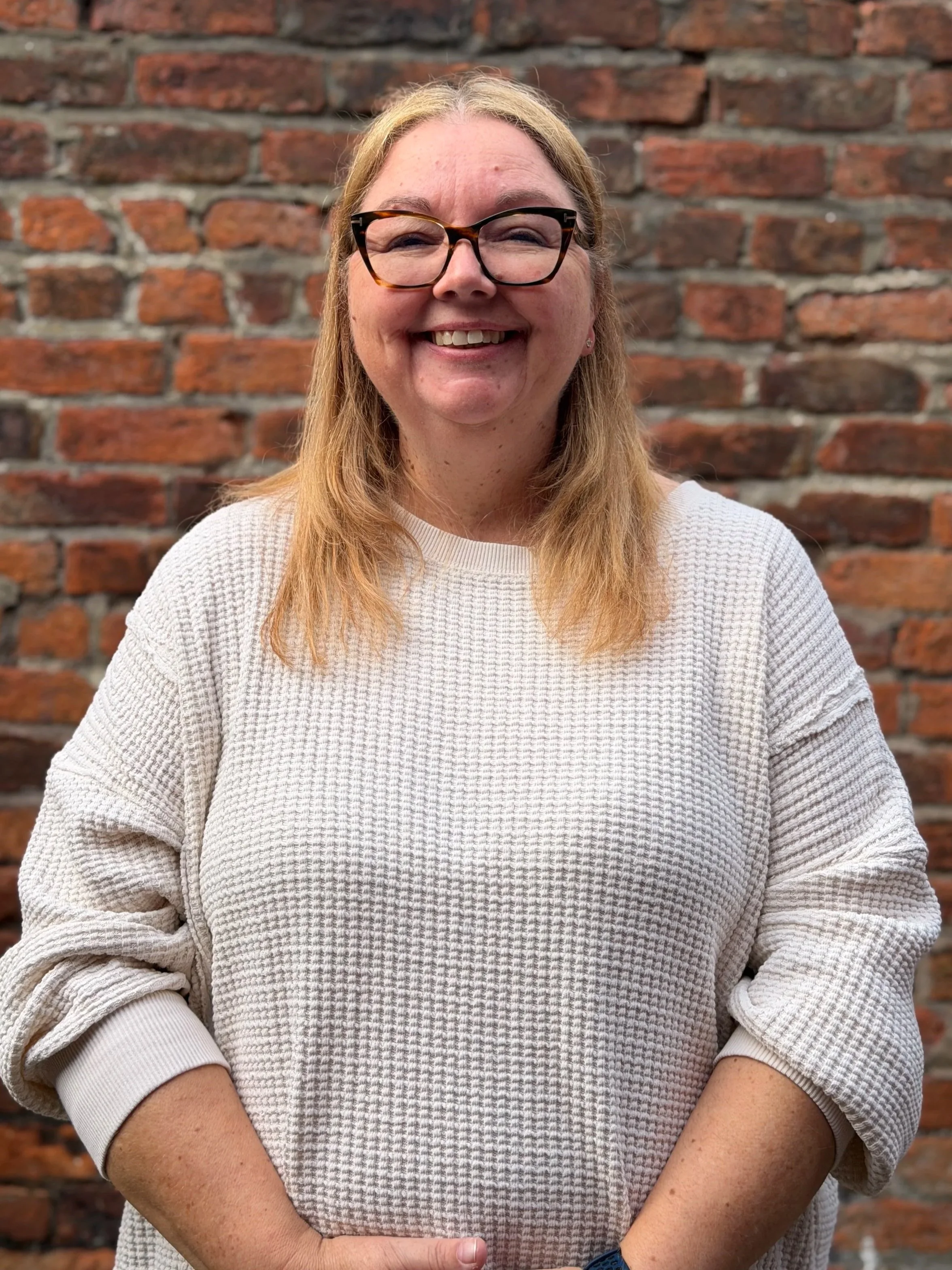 Julie - A woman with blonde hair and glasses smiling while sitting in front of a red brick wall.
