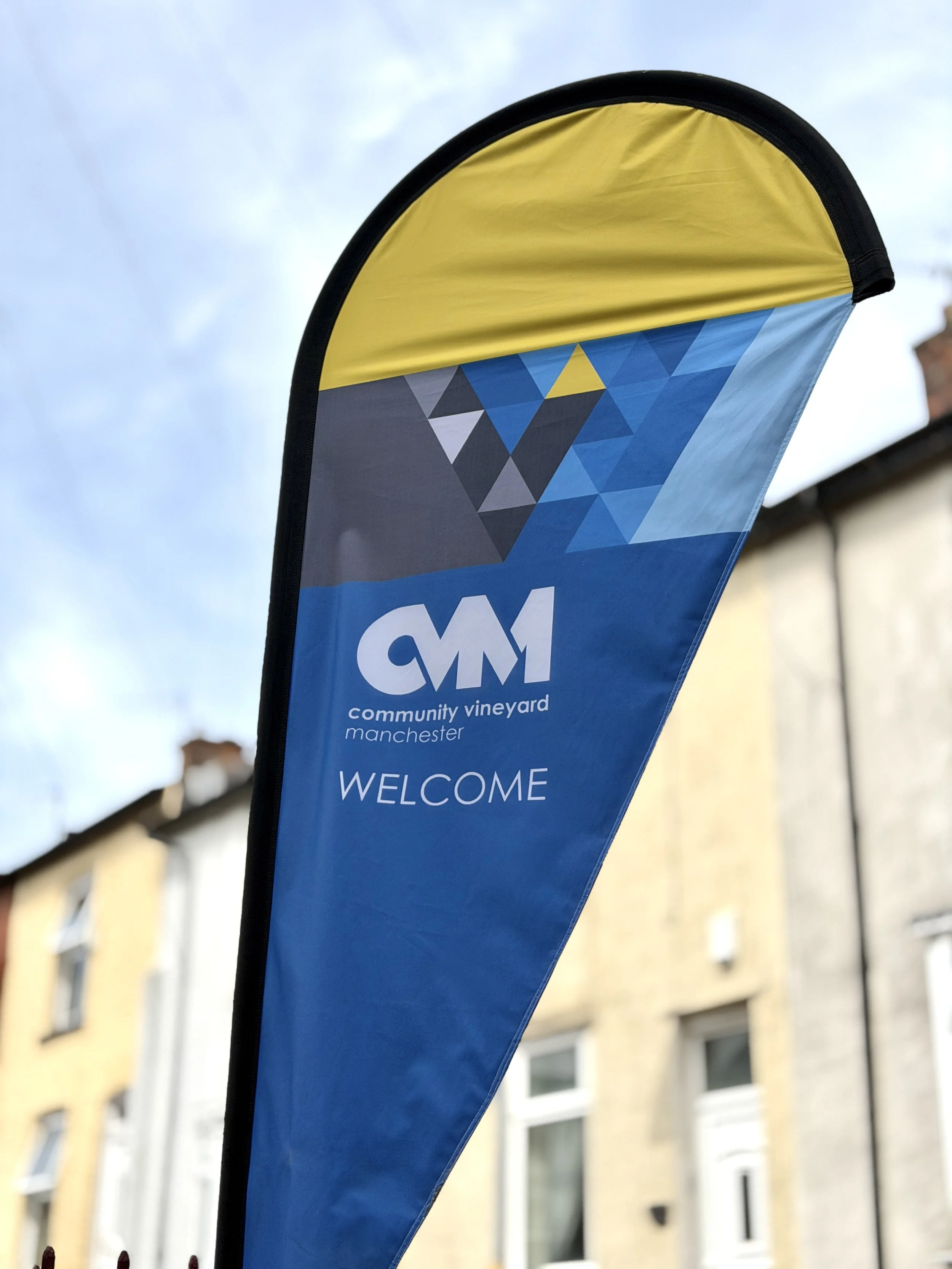 Blue and yellow feather-shaped banner with the text 'Community Vineyard Manchester' and a geometric logo, set against a background of buildings and a cloudy sky.