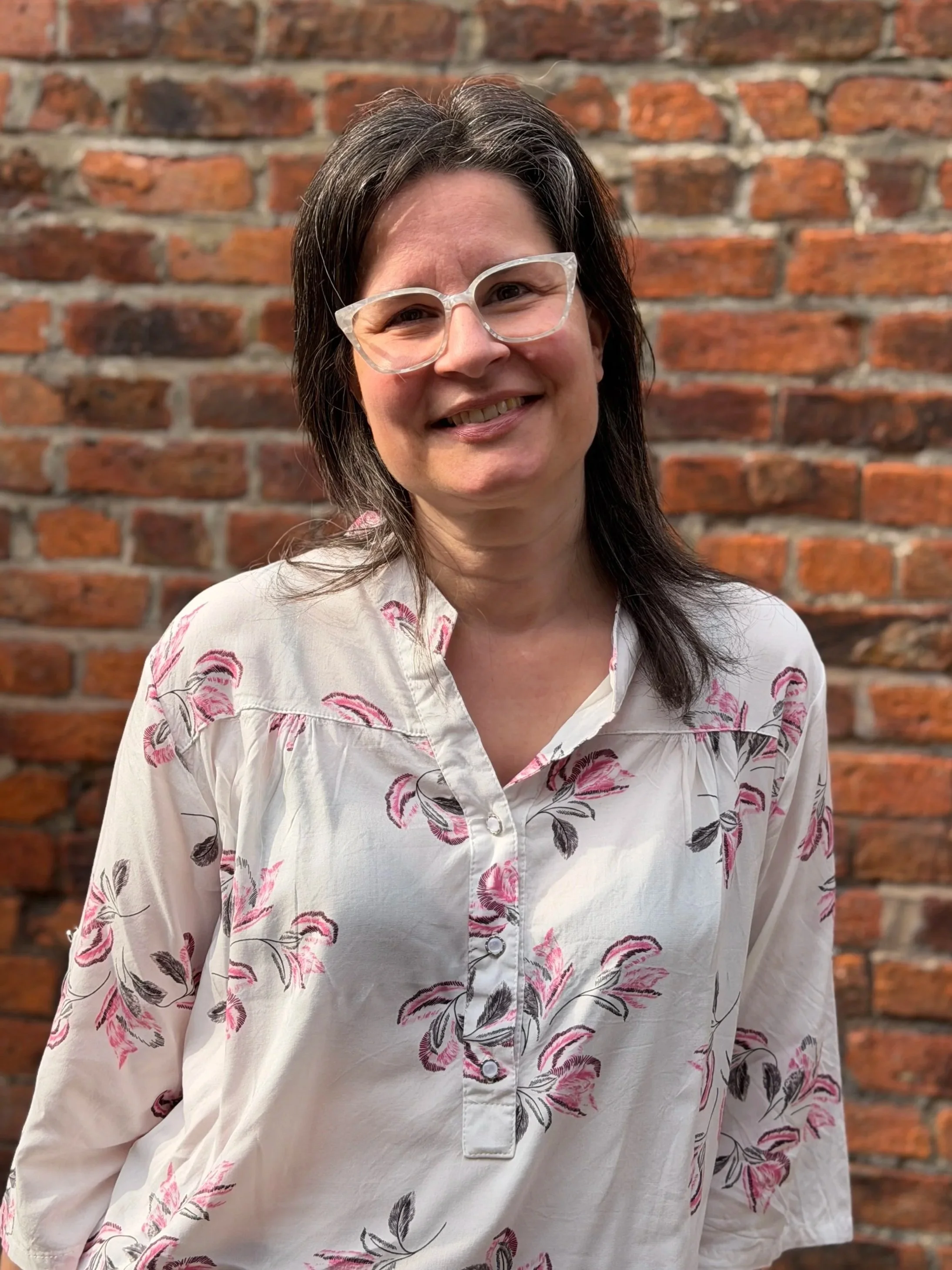 Edina - A woman with dark brown hair, wearing glasses with a clear frame, smiling and standing in front of a brick wall. She is wearing a white shirt with a pink and black floral pattern.
