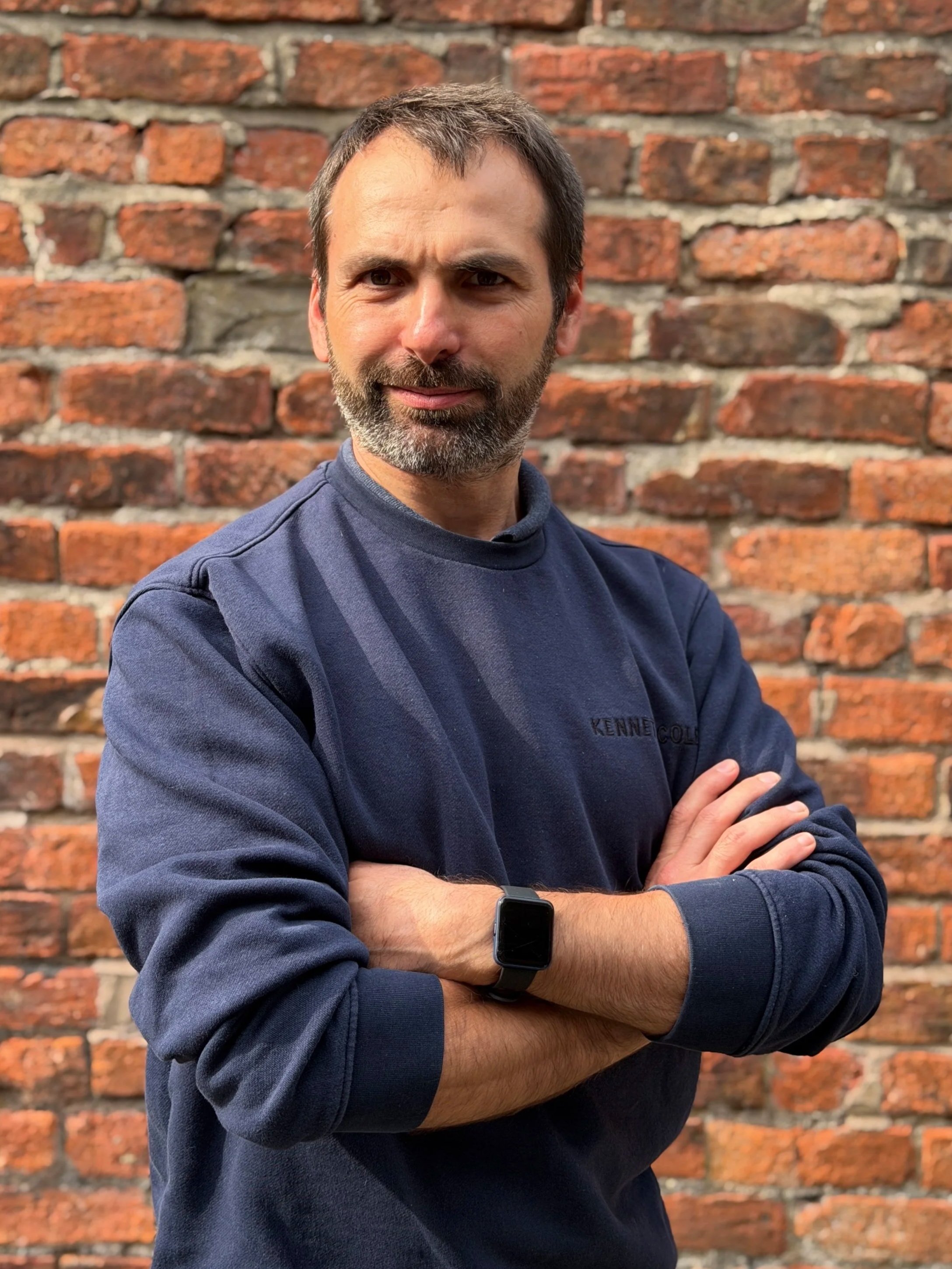 Josh - A man with brown hair and a beard stands in front of a red brick wall. He is crossing his arms and wearing a dark blue sweatshirt. He has a black smartwatch on his left wrist.