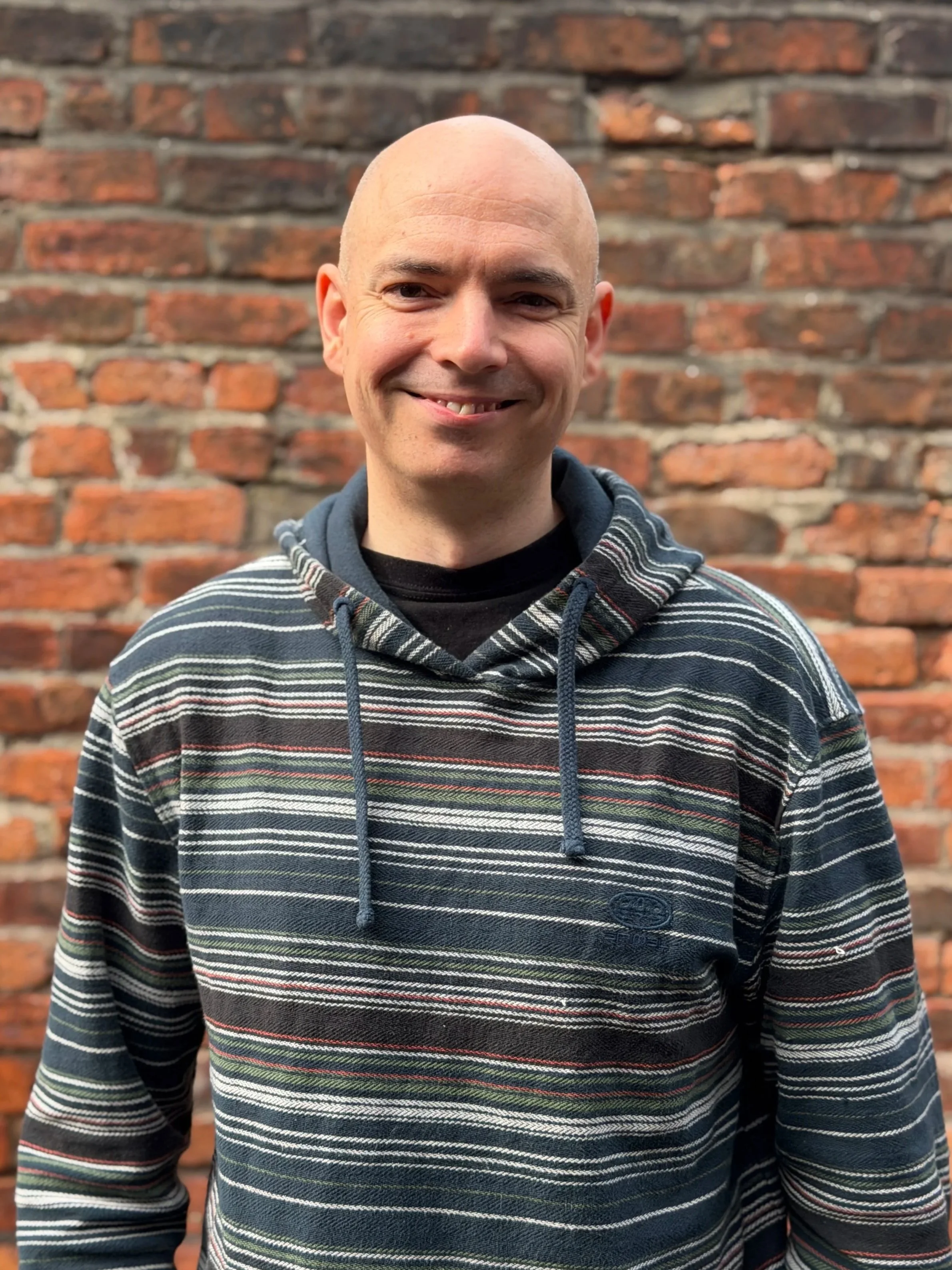 Ben - A man with a shaved head and a striped hoodie smiling in front of a brick wall.