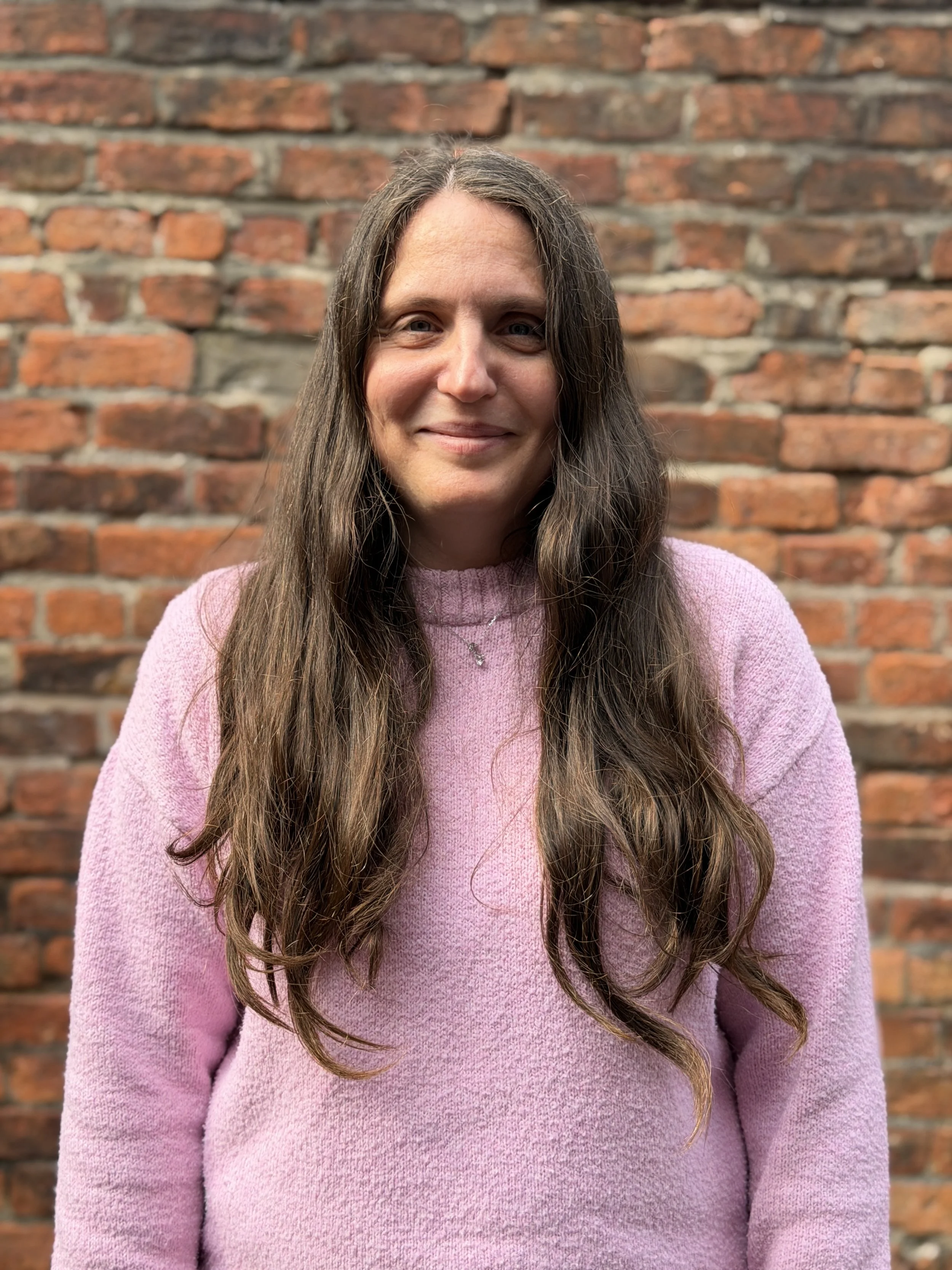 Lucy - A woman with long brown hair wearing a pink sweater standing in front of a brick wall.
